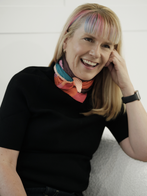 A woman with blonde hair and colorful streaks in her bangs, smiling and wearing a black top and a colorful scarf, sitting indoors against a white wall.