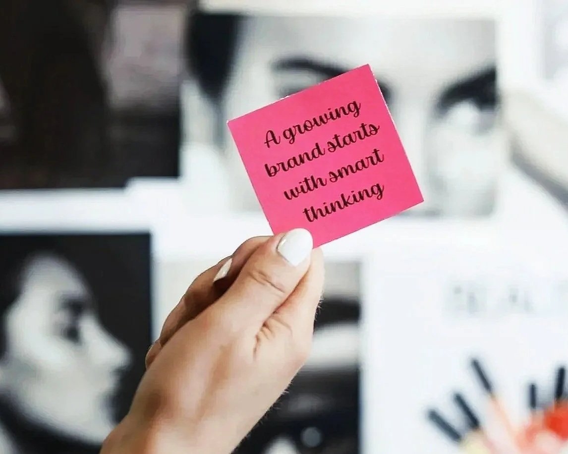 A hand holding a pink note card with the text "A growing brand starts with smart thinking" in black cursive font, blurred black and white artwork in the background.