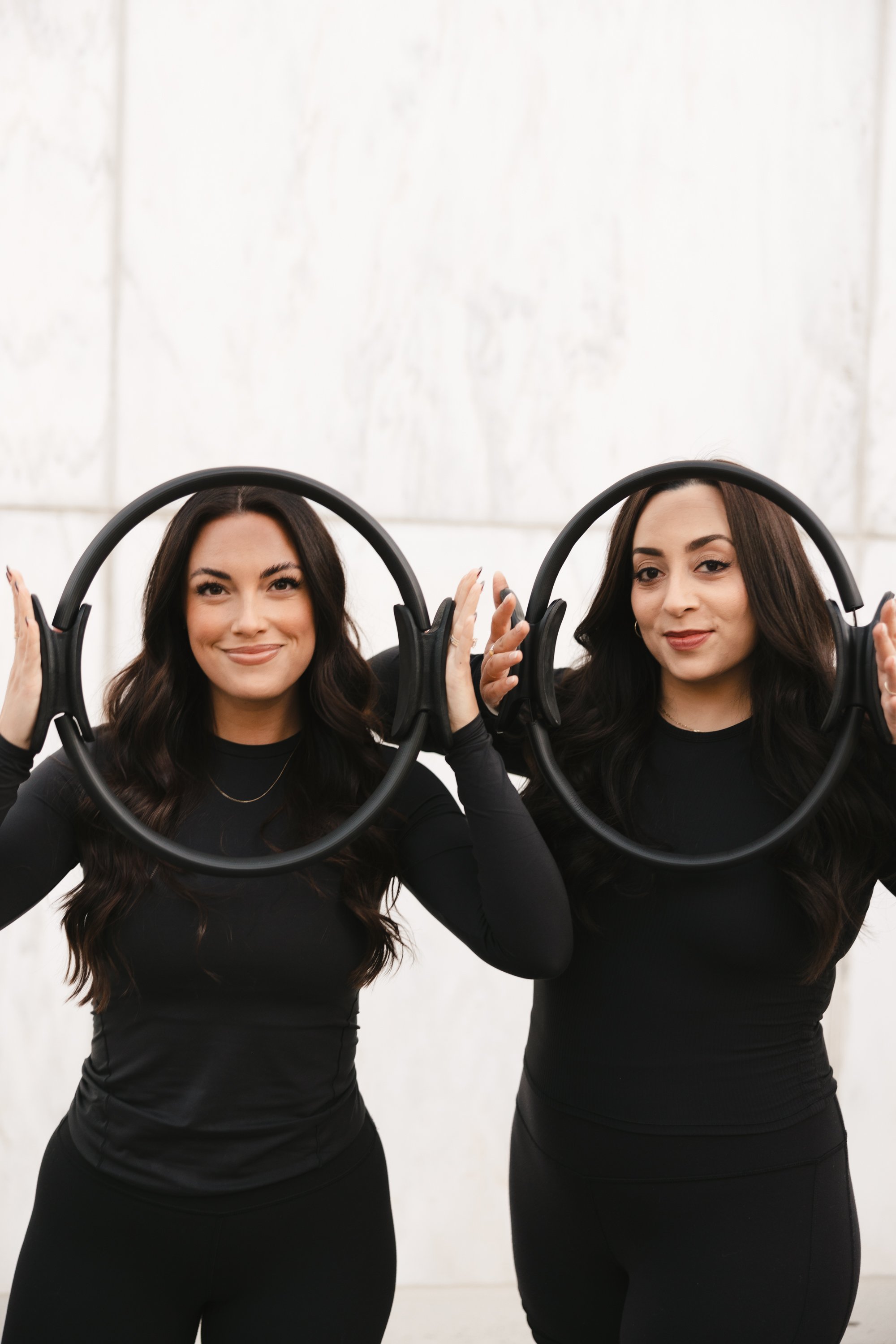 Two women standing side by side, holding black headphones around their necks, smiling, and wearing black long-sleeve tops and black skirts, against a white background.