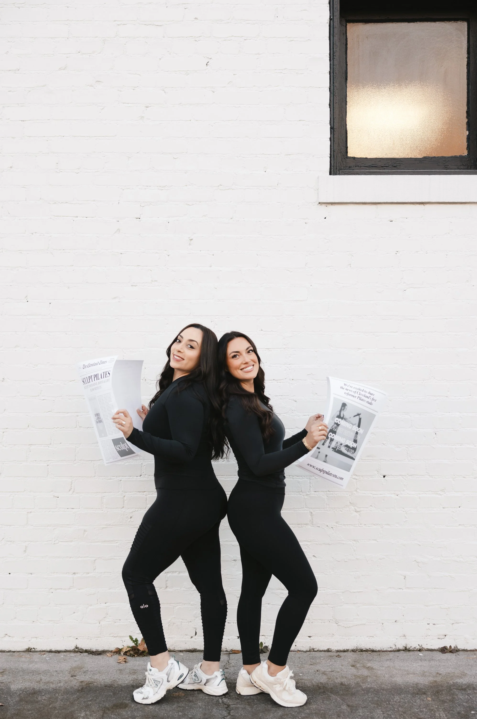 Two women with long dark hair, dressed in black workout attire, are standing back-to-back in front of a white brick wall. They are both holding newspapers and have playful, smiling expressions.
