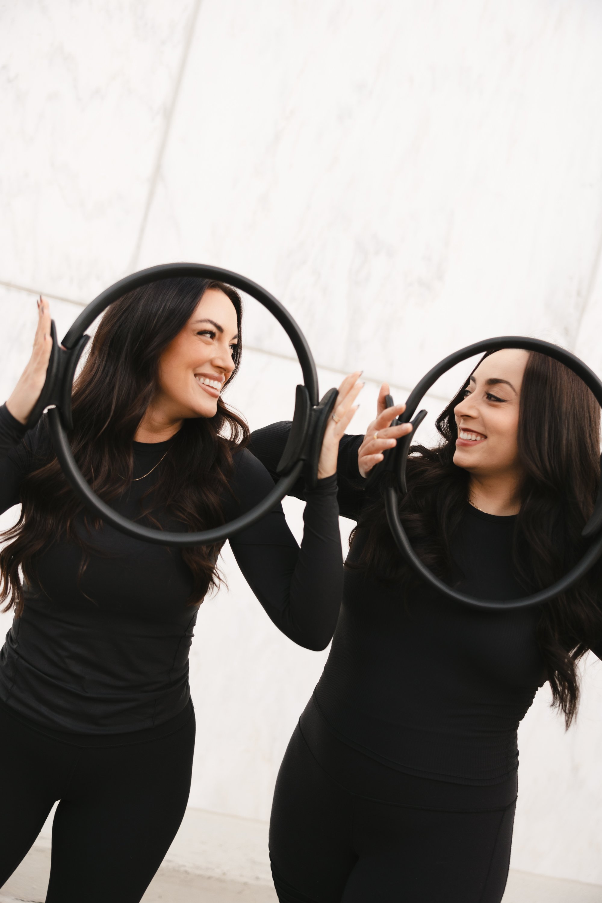 Two women smiling and touching mirrors in an indoor setting, both wearing black long-sleeve shirts and black pants.