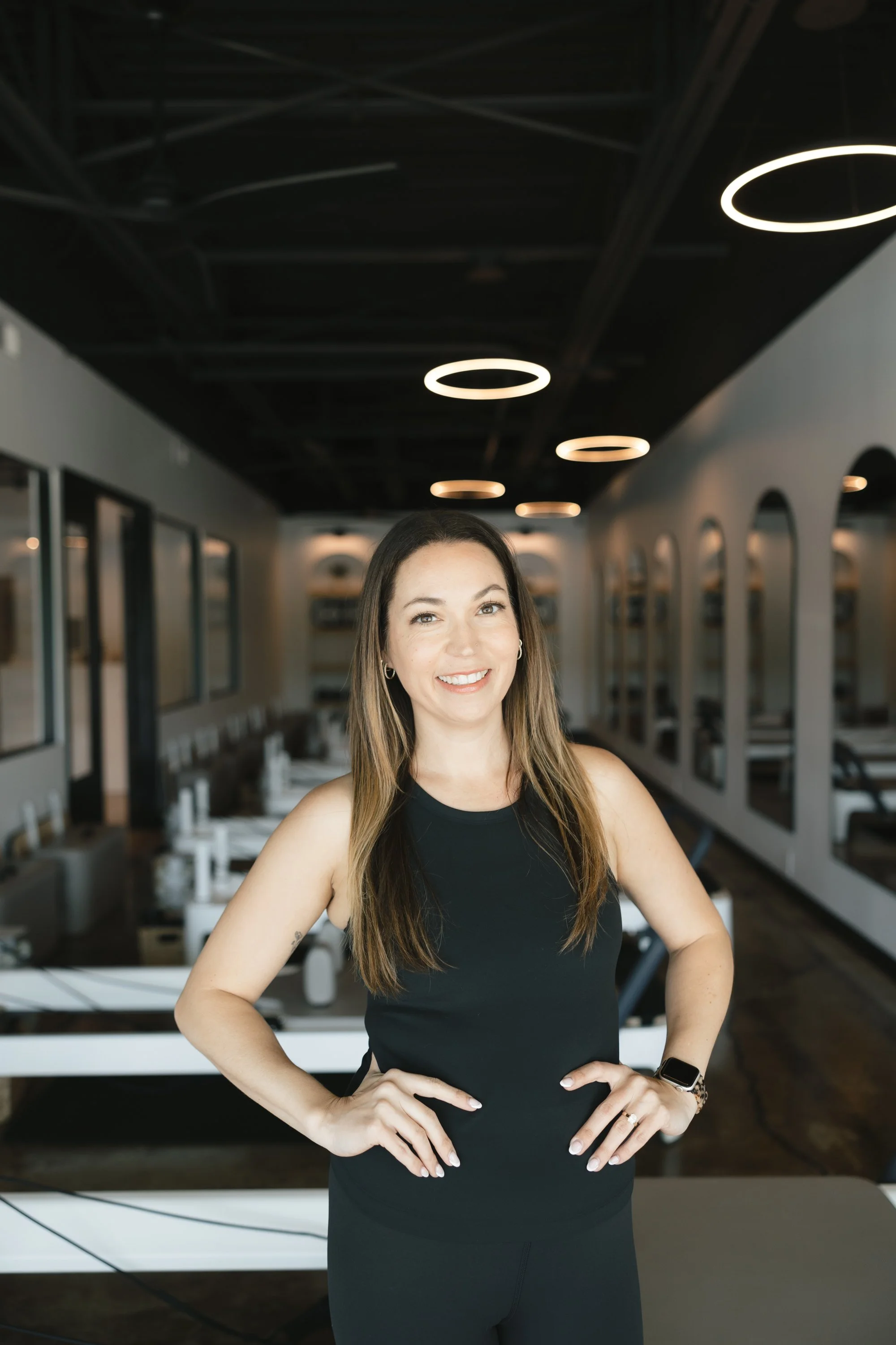 A woman with long brown hair, wearing a black sleeveless top, standing in a modern, well-lit room with circular ceiling lights, windows on the side walls, and a row of white chairs and tables in the background.
