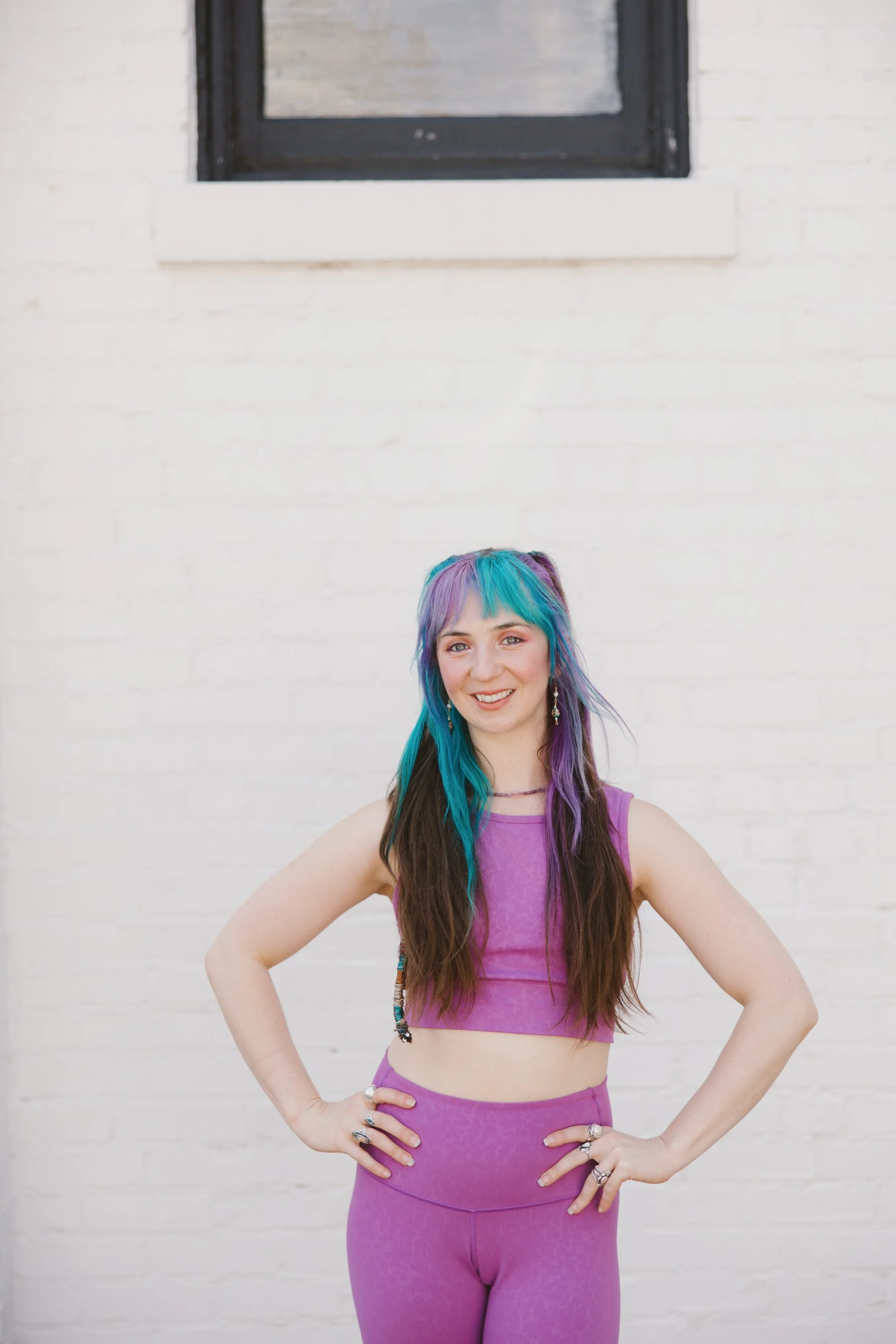 A young woman with long brown hair and colorful hair clips, wearing a pink sports bra and matching leggings, standing with her hands on her hips against a white brick wall with a window above.