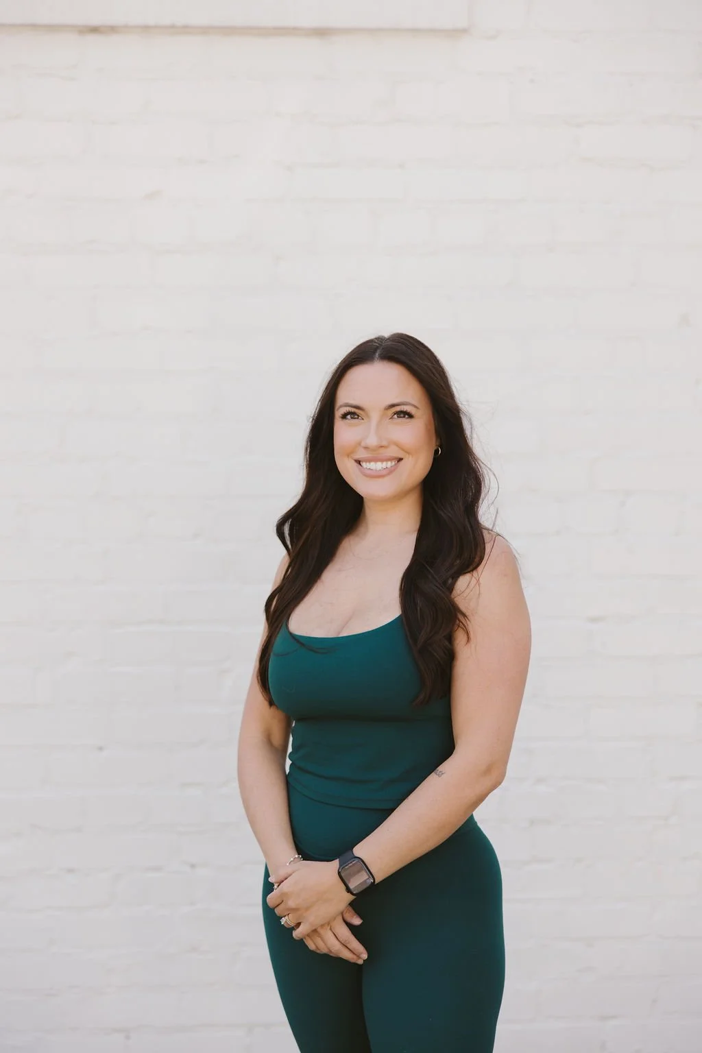 A woman with long dark hair smiling, wearing a teal sleeveless top and matching pants, stands against a white brick wall.