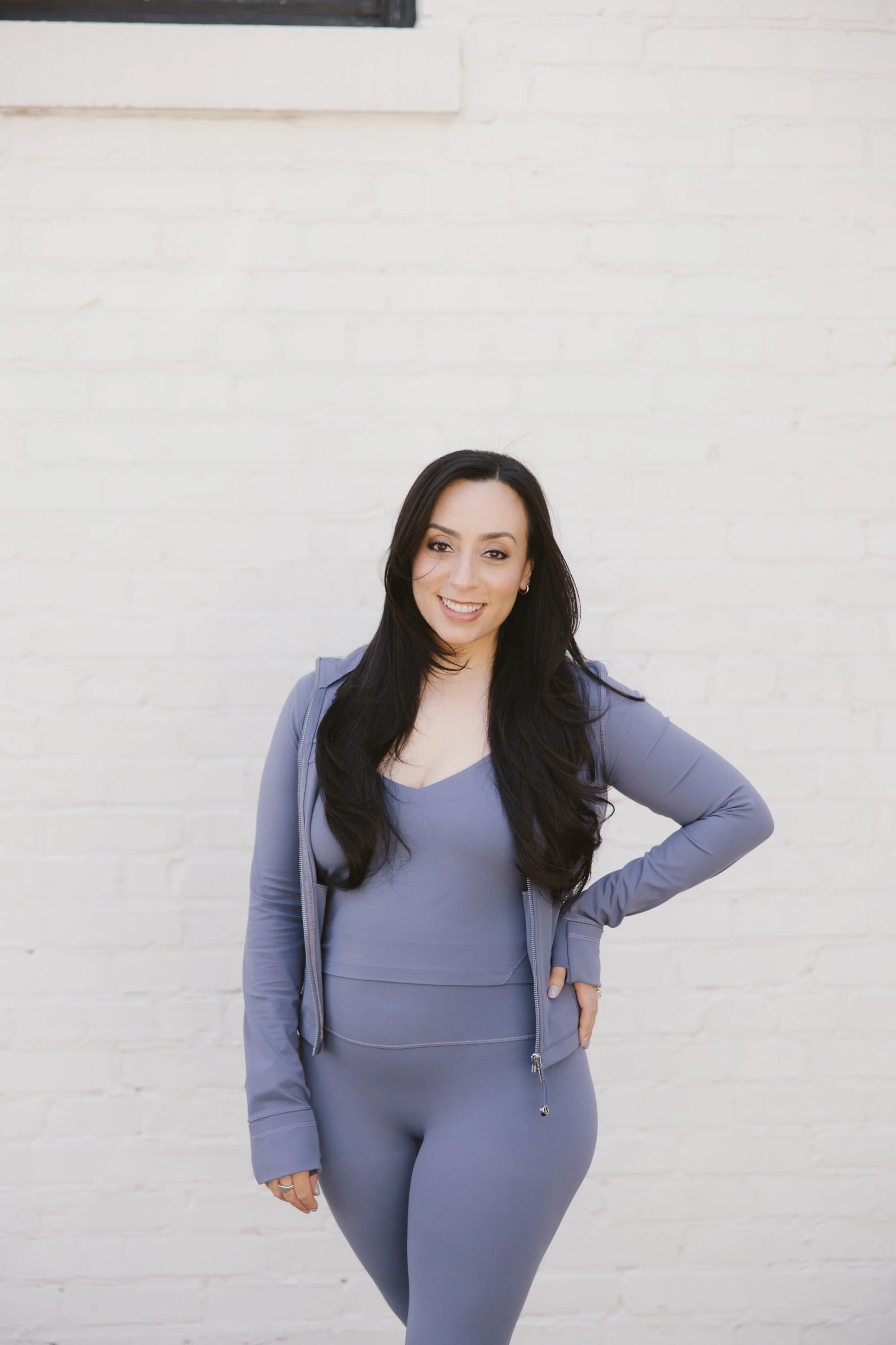 A woman with long dark hair wearing a matching light purple workout outfit and jacket, standing against a white brick wall, smiling with her hand on her hip.