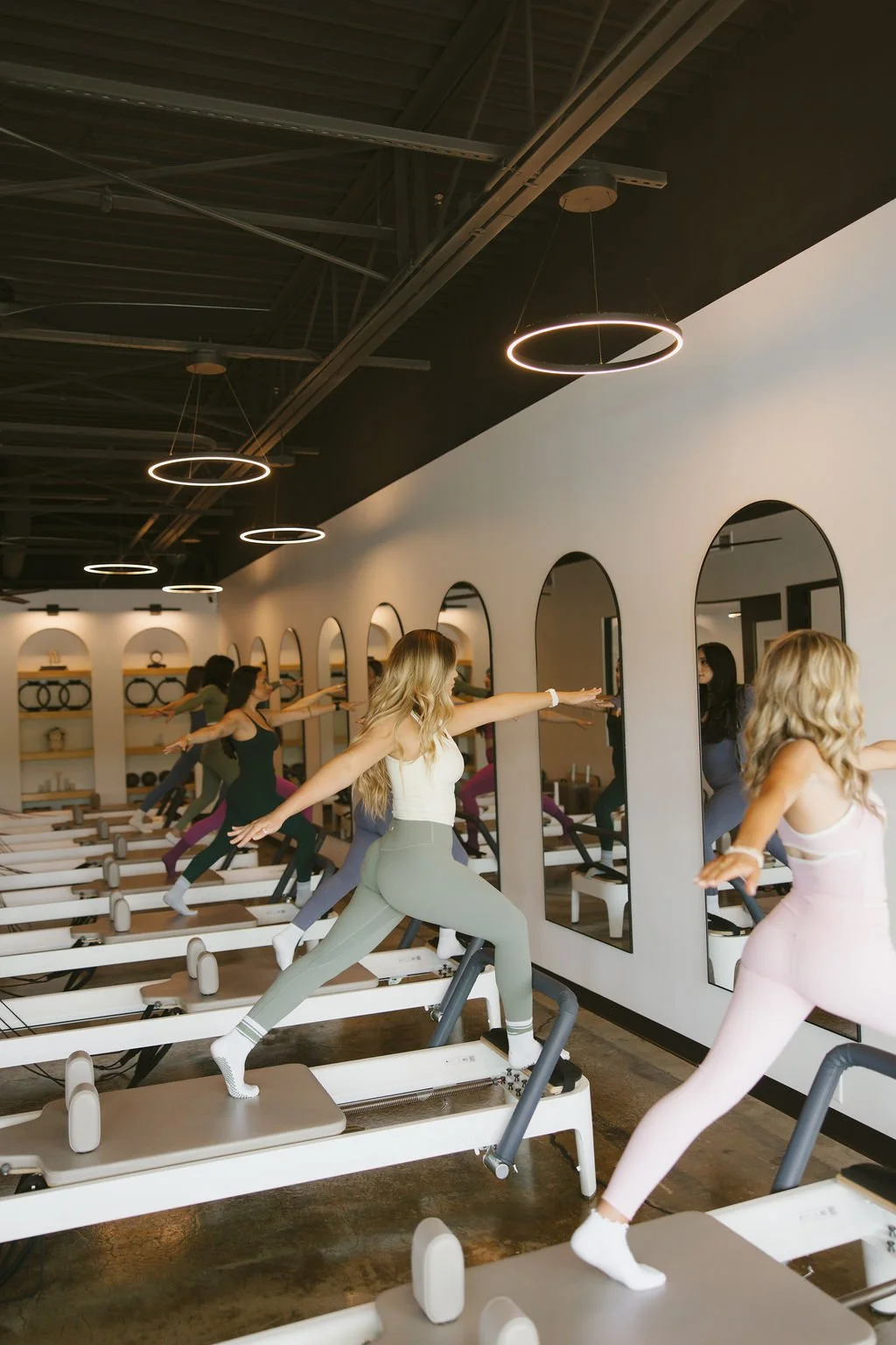 A group of women in workout clothes participating in a fitness class on reformer Pilates machines in a modern studio with arches and round mirrors on the wall.
