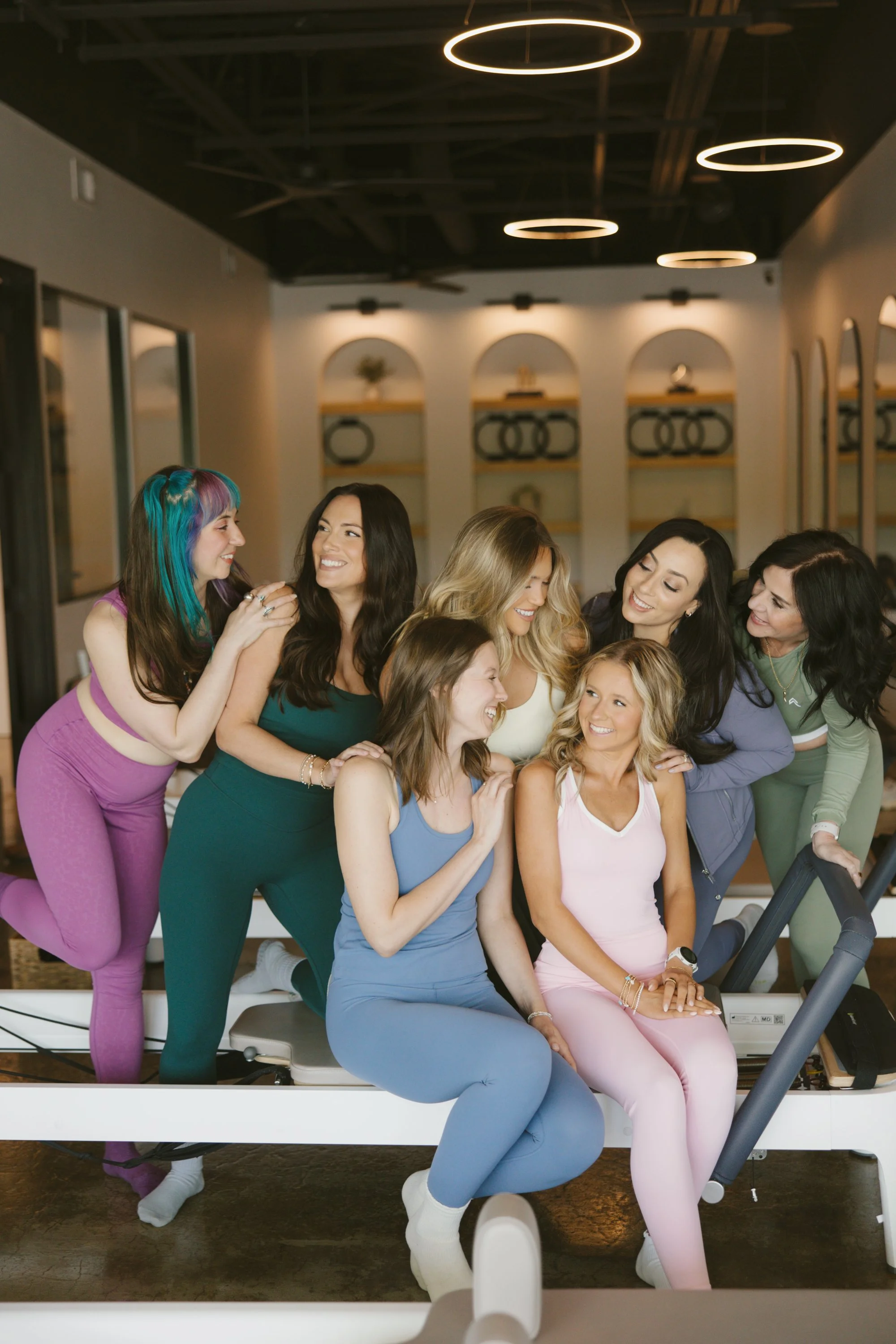 A group of women on a treadmill at a gym, smiling and interacting with each other, dressed in colorful workout clothes.