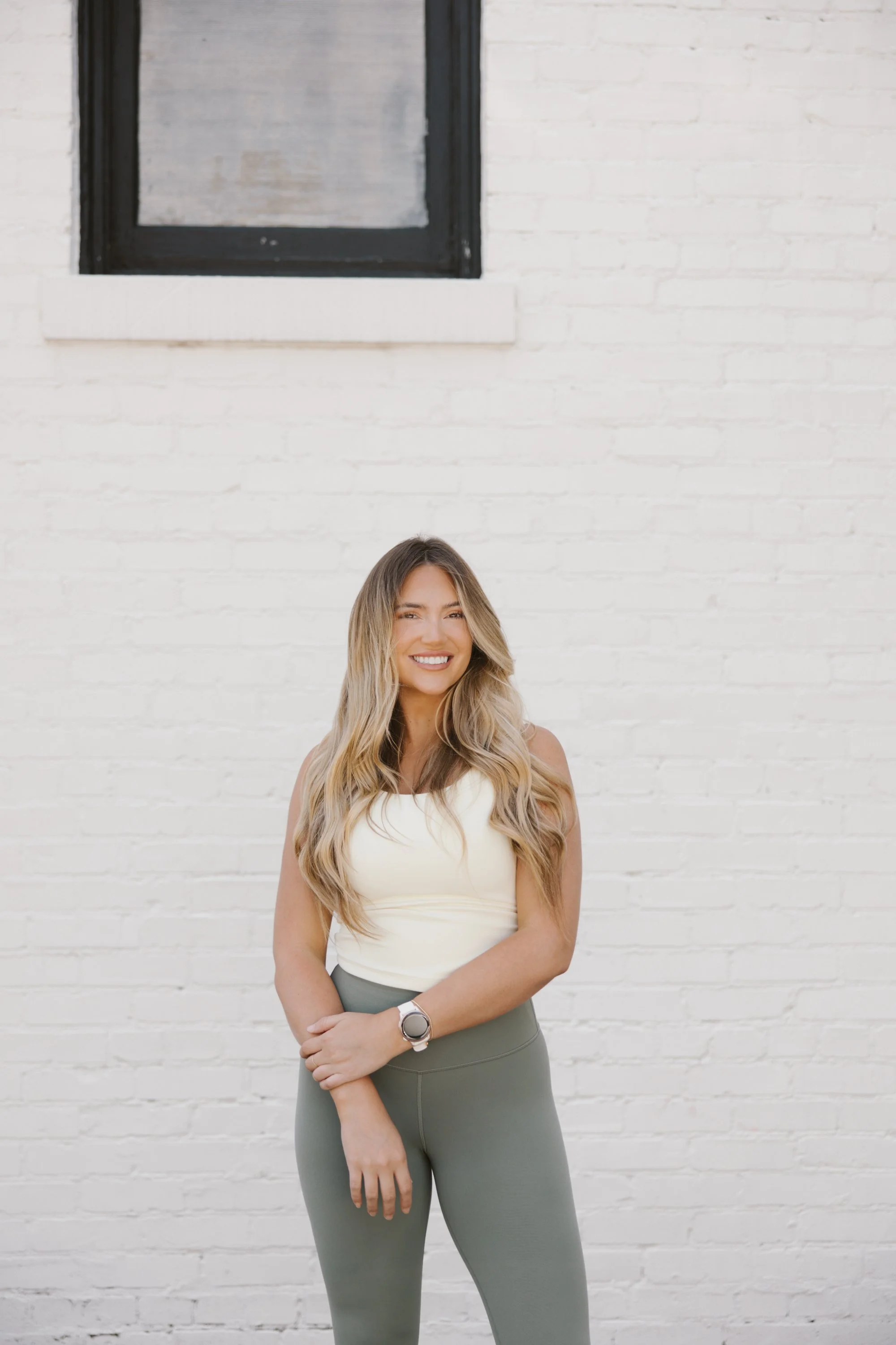 A woman with long wavy blonde hair smiling, wearing a light-colored sleeveless top and high-waisted green athletic leggings, standing in front of a white brick wall with a black-framed window.