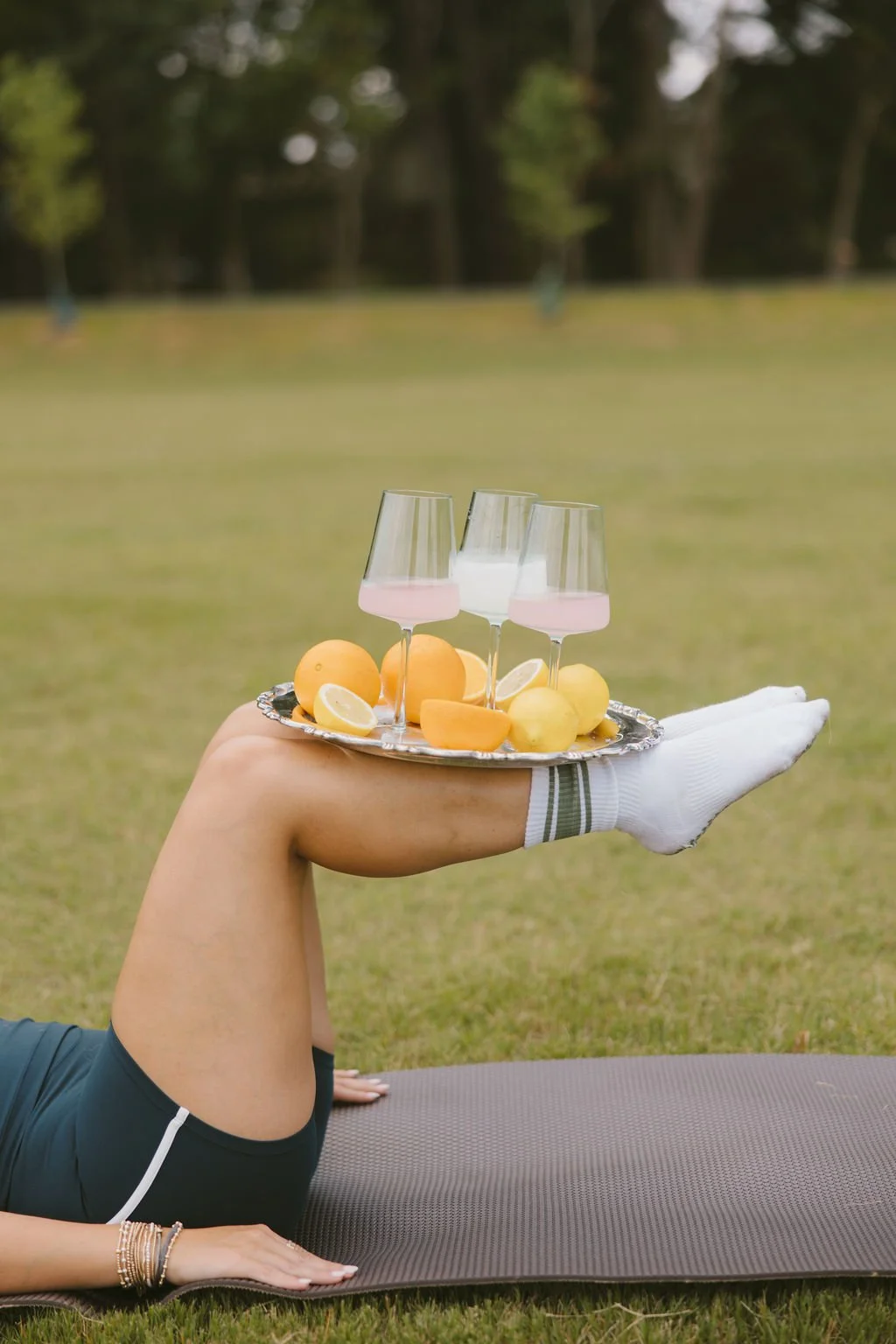 A person lying on a yoga mat outdoors, holding a silver platter with three wine glasses filled with pink and white drinks, lemon wedges, and yellow and orange fruits, with a background of grass and trees.
