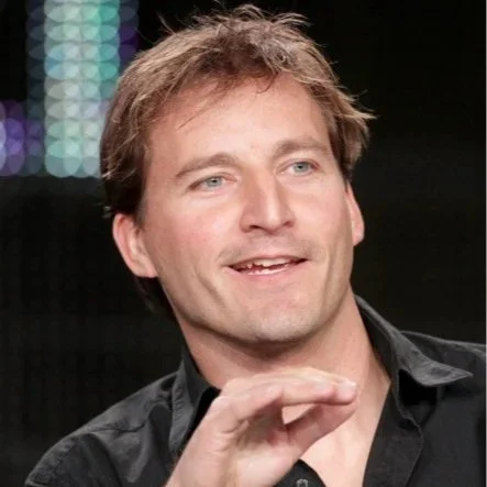 A man with light brown hair and blue eyes, smiling, wearing a black collared shirt, against a dark background with colorful lights.