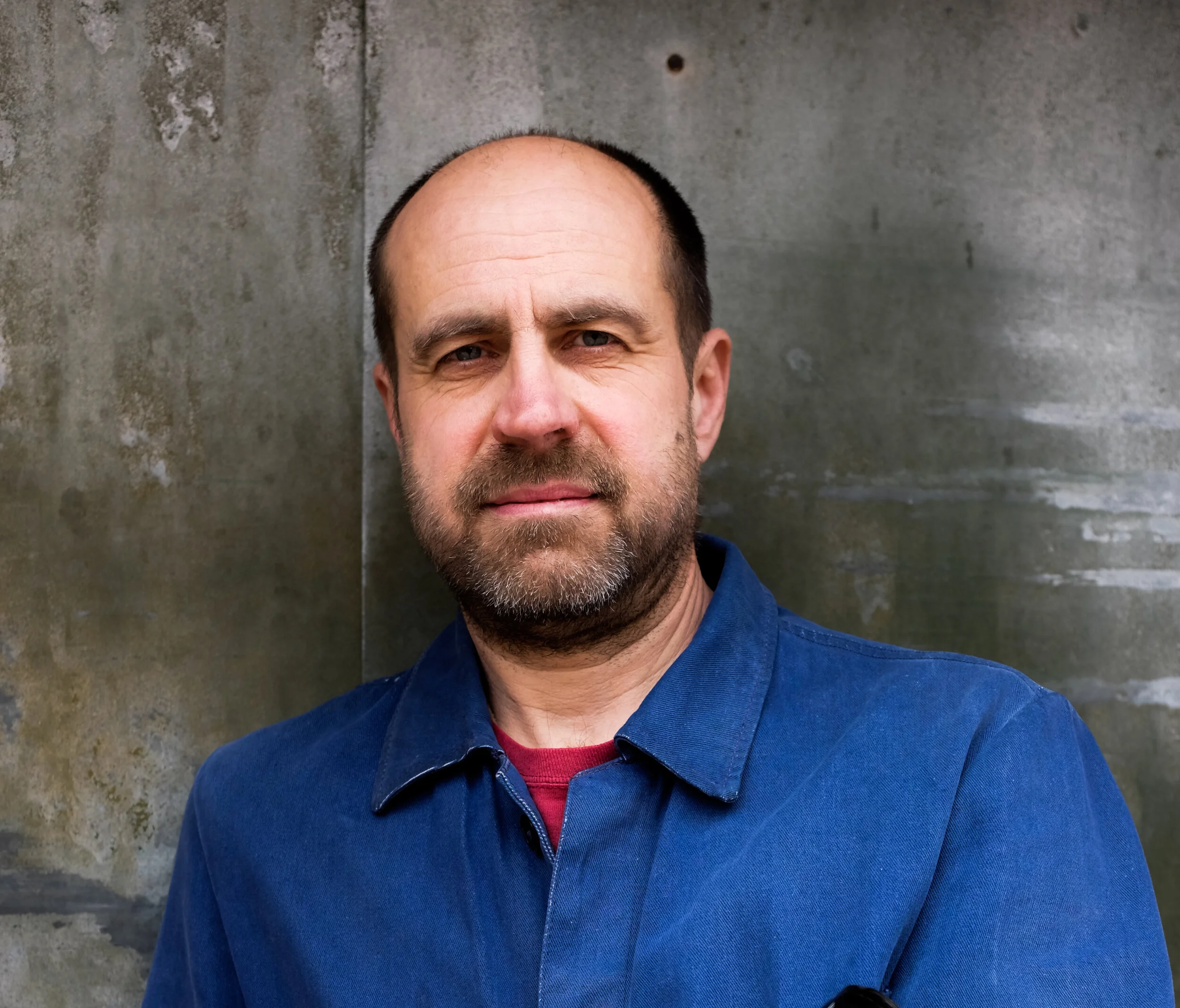 A man with a shaved head and beard is standing outdoors against a gray concrete wall, wearing a dark blue shirt and a red shirt underneath.
