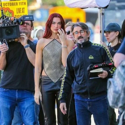 A woman with long red hair and a woman wearing sunglasses and a black cap sit in the middle of a filming crew outdoors, with cameras and crew members around them and a yellow sign in the background.