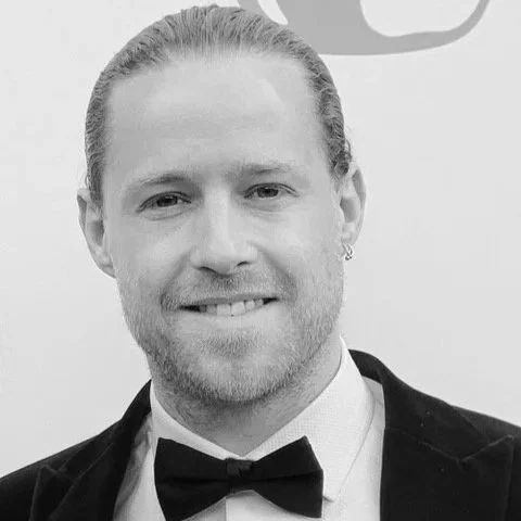 Portrait of a man with long hair tied back, a beard, dressed in a tuxedo with a bowtie, smiling in front of a plain background.