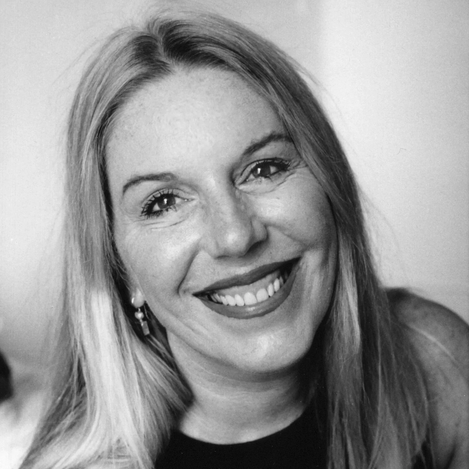 Black and white portrait of a smiling woman with long hair, earrings, and wearing a dark top.
