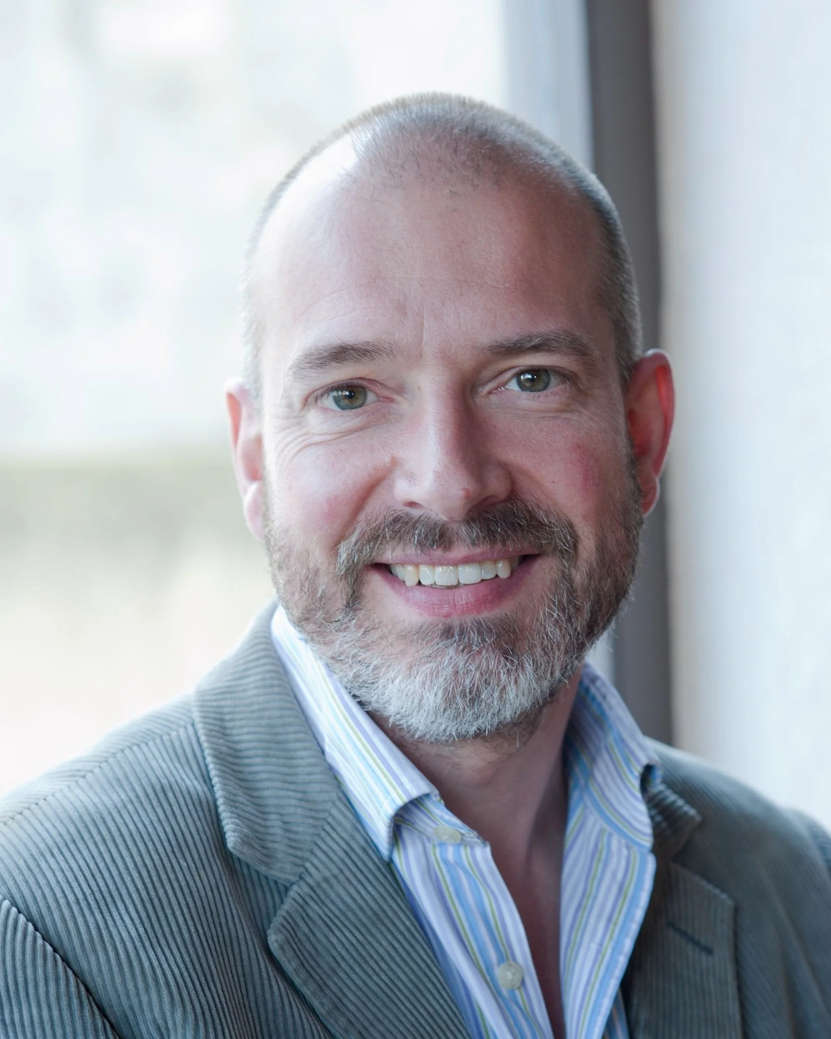 Close-up of a smiling man with a beard, wearing a striped collared shirt and a light-colored blazer, standing indoors near a window.