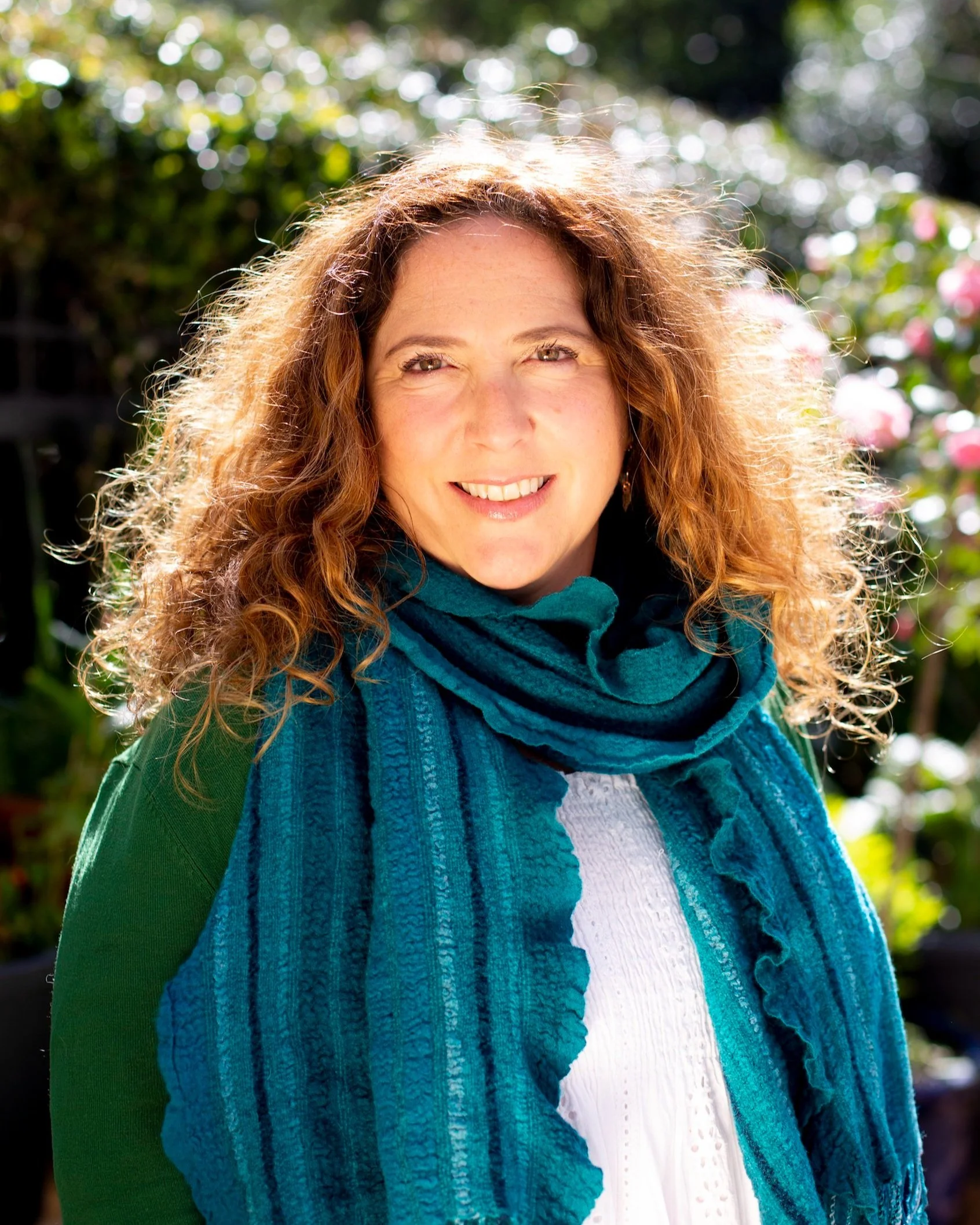 A woman with curly red hair smiling outdoors, wearing a teal blue scarf, a white top, and a green jacket, with a background of green foliage and pink flowers.