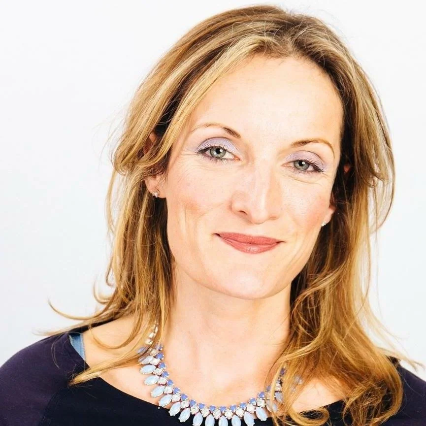 A woman with light brown wavy hair, wearing a dark top and a statement necklace, smiling softly against a plain light background.
