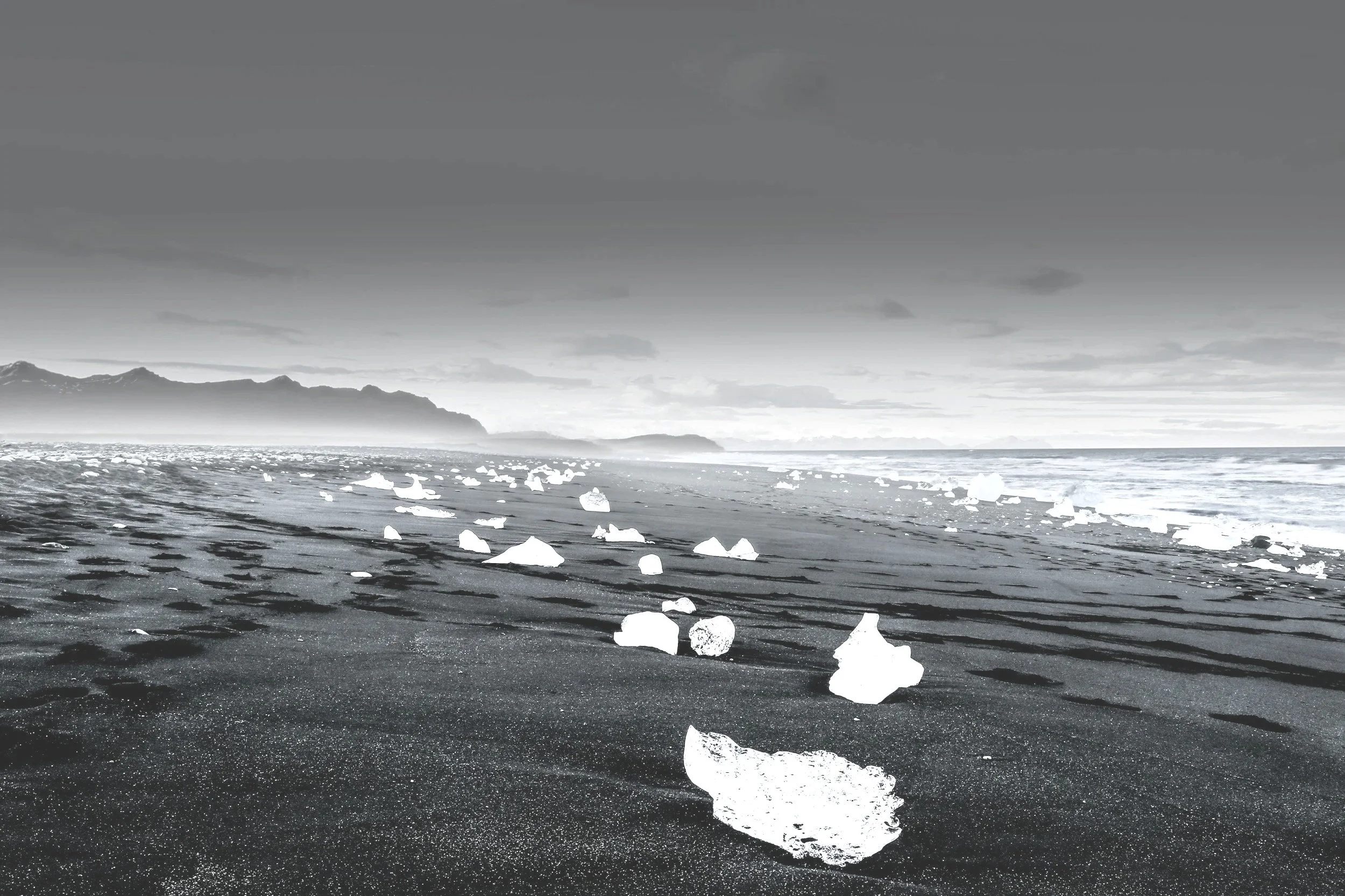 A black and white photo of a beach with ice chunks scattered along the shoreline and mountains in the background under a cloudy sky.