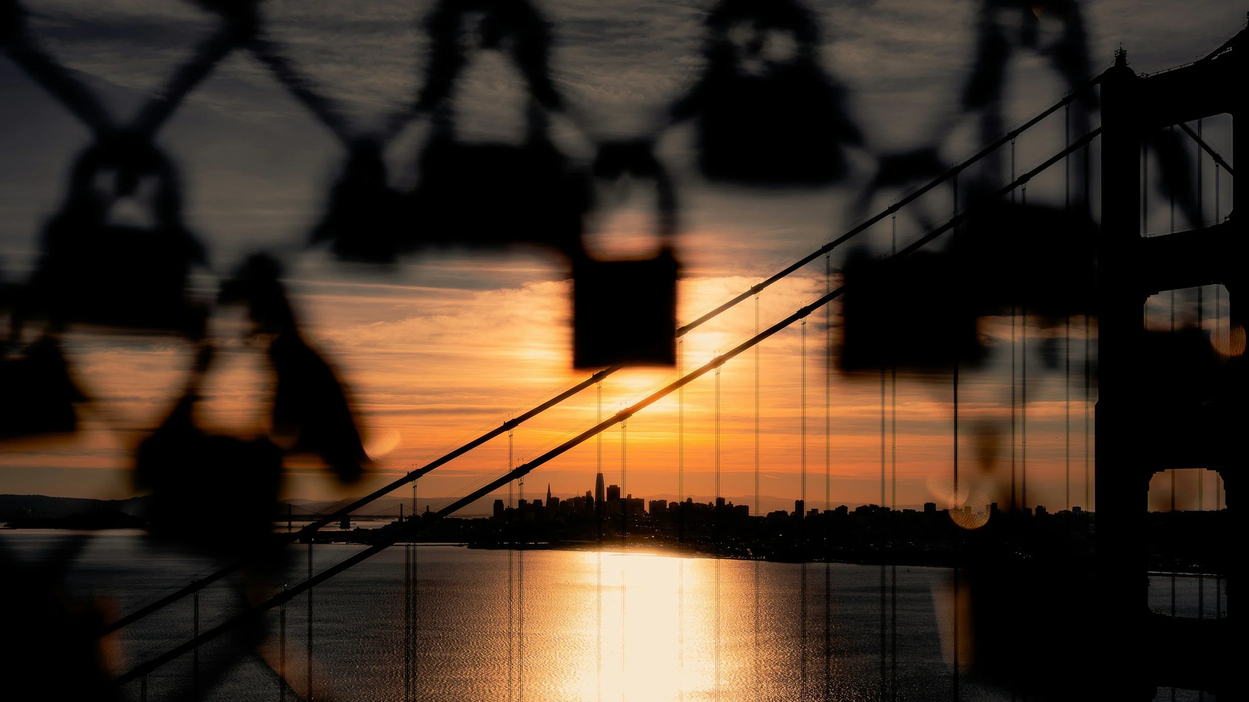 Silhouettes of a chain-link fence in the foreground with a city skyline and a bridge in the distance at sunset, with the sun partially obscured.