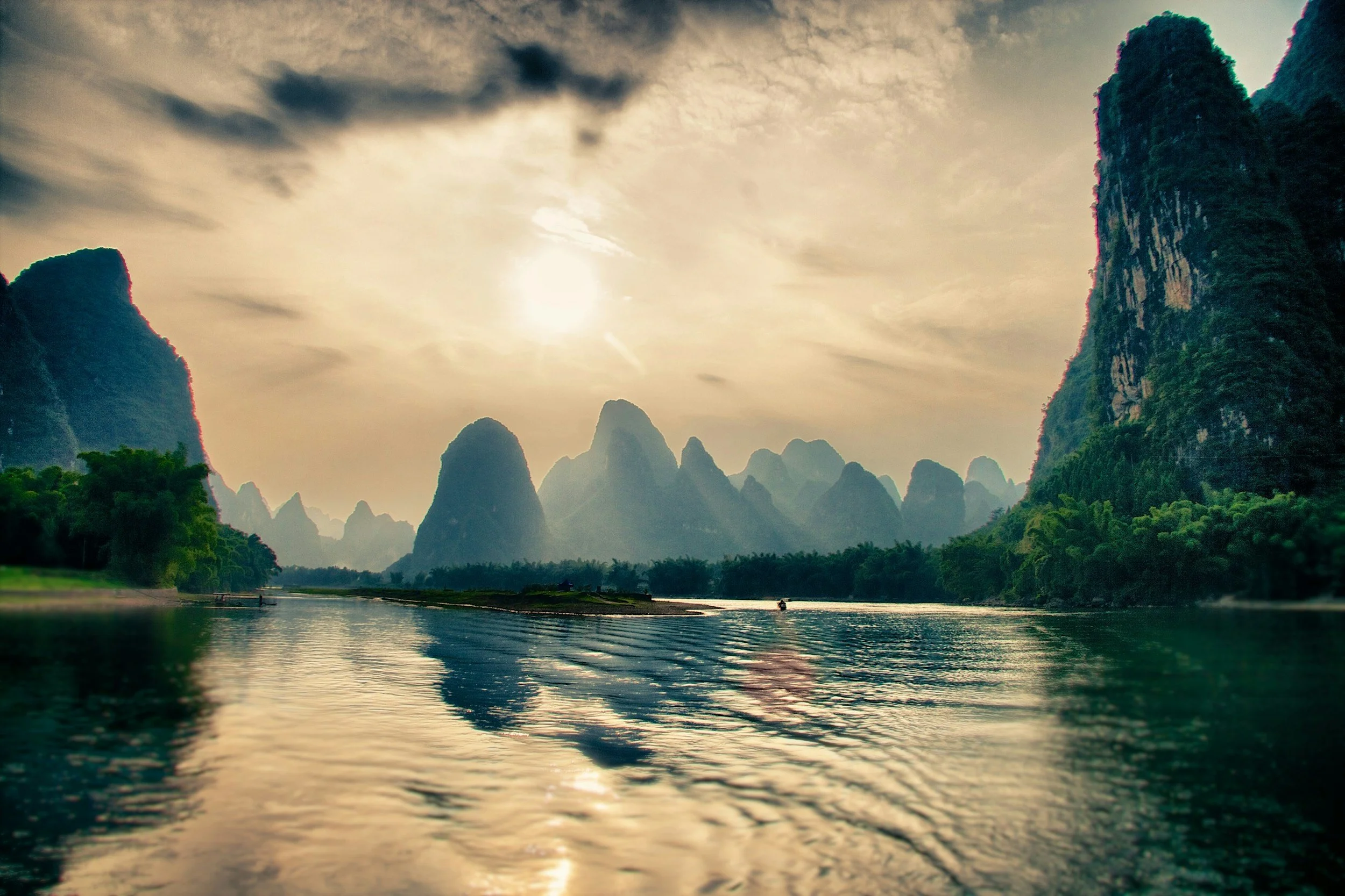A scenic river flowing through lush green valleys surrounded by tall karst mountains with cloudy sky and the sun partially obscured.