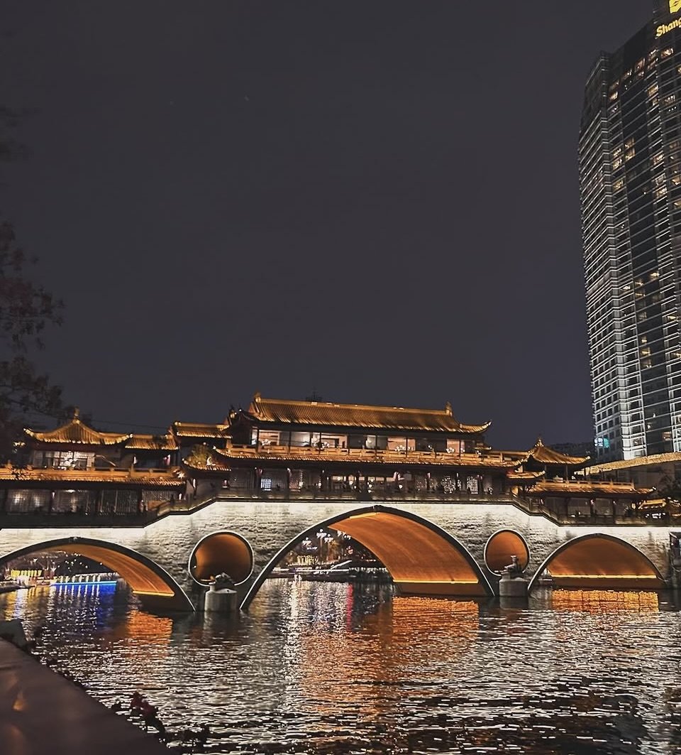 Night view of a traditional Chinese-style bridge with illuminated rooftops and arches over a river, with modern skyscrapers in the background.