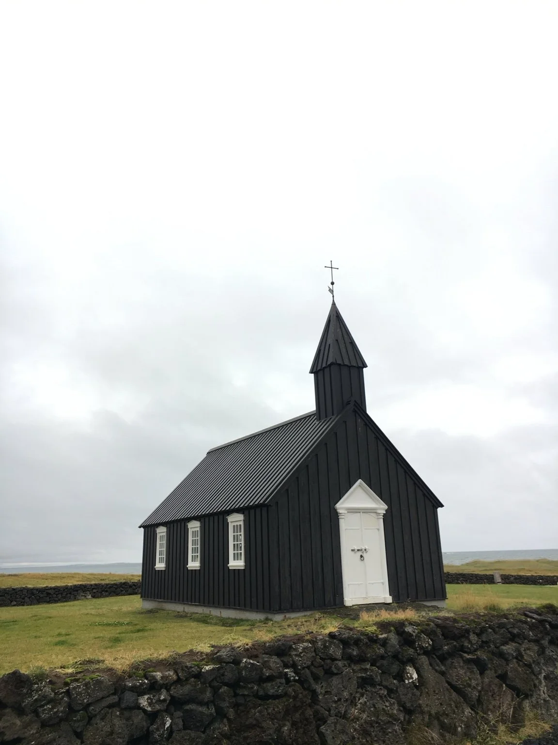 Small black church with white door and trim, situated on grassy area with stone wall in foreground, overcast sky in background.