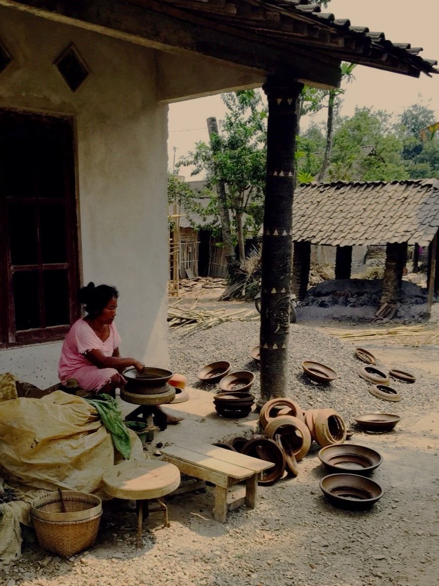 A woman in a pink dress sitting outside a house, preparing clay pots in a rural village setting with more clay pots scattered around, some stacked and some on the ground.