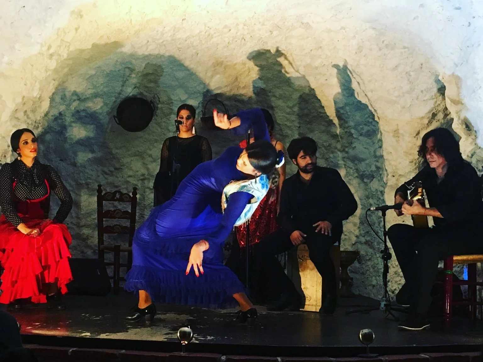 A flamenco dance performance with a singer and guitarist in a cave-like venue. The female dancer in a blue dress is bowing, while the seated guitarist is playing. Other women in red and black are seated or standing, watching the performance.