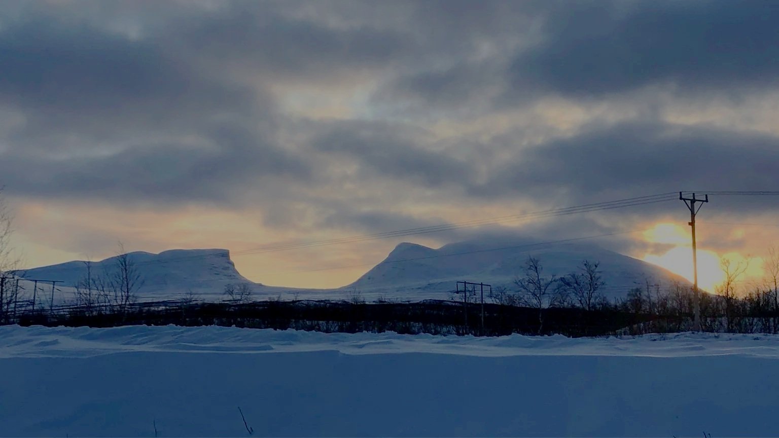 Snow-covered landscape with mountains in the distance, power lines, leafless trees, and a cloudy sky during sunset or sunrise.