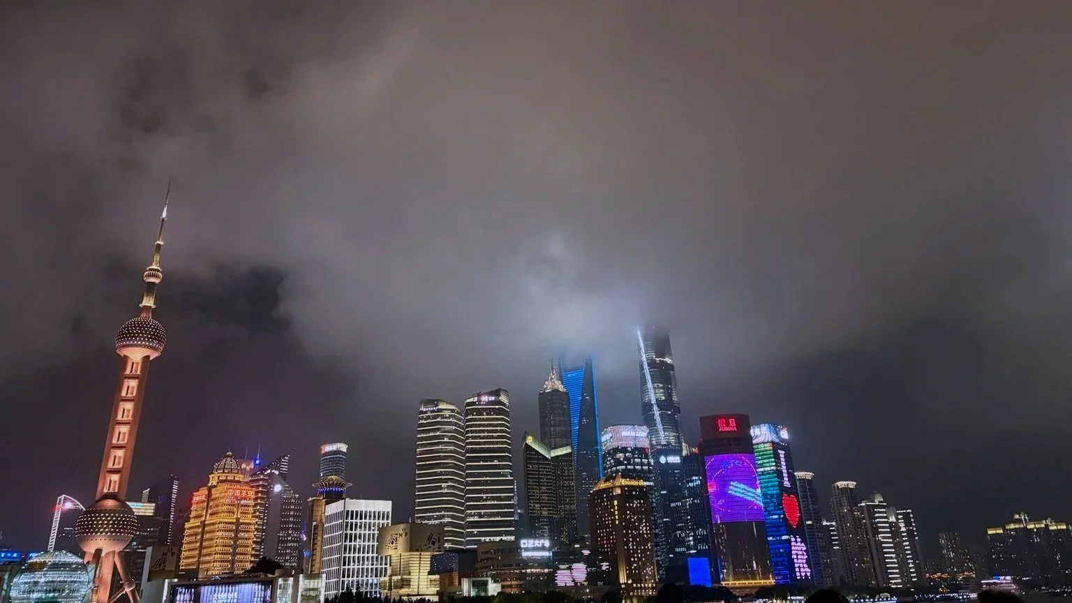 Night view of Shanghai skyline with illuminated skyscrapers, including the Oriental Pearl Tower, under cloudy skies.