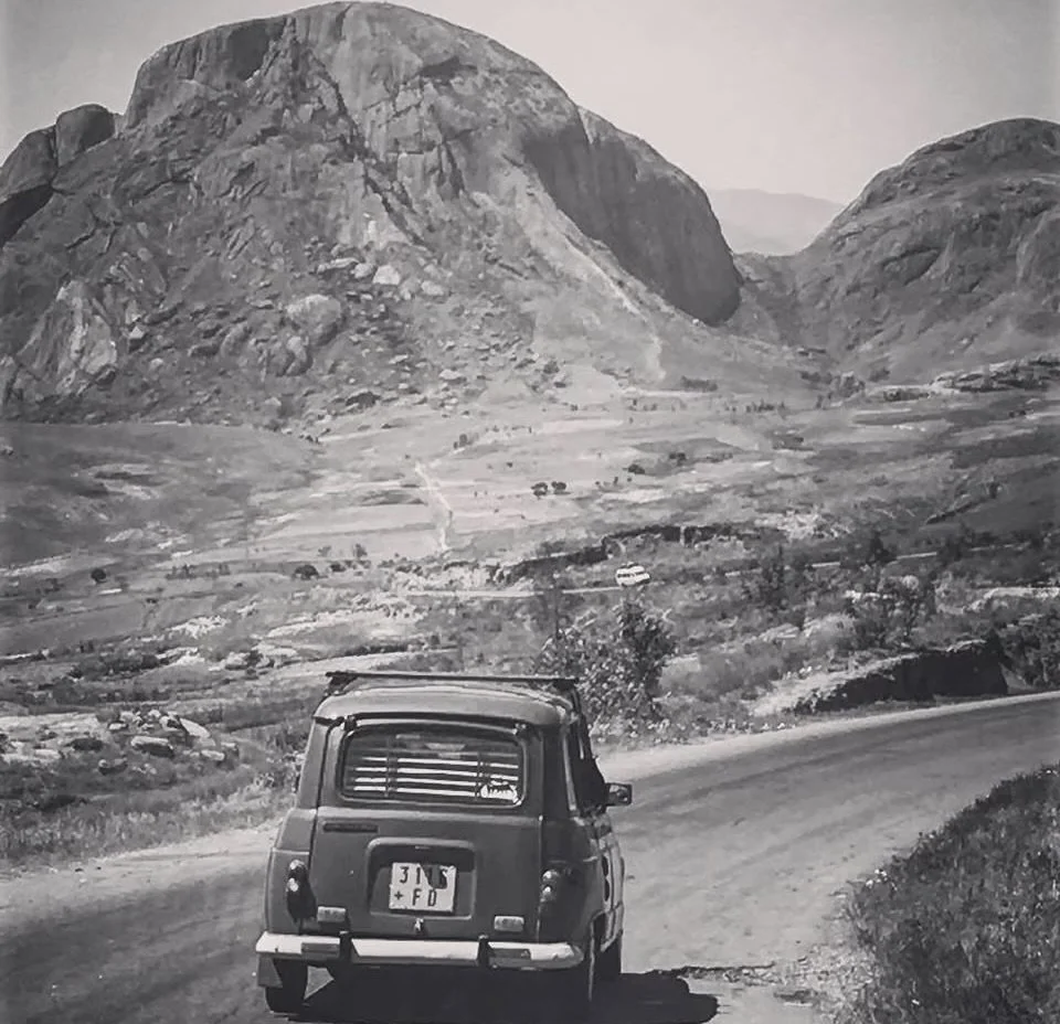 A vintage car driving along a winding mountain road with rocky hills and mountains in the background.