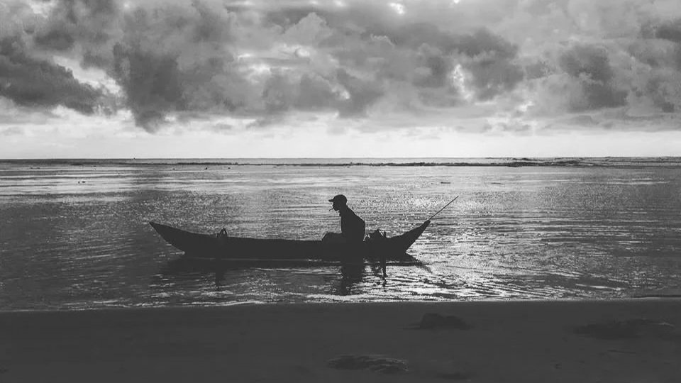 Silhouette of a person in a small boat on a calm body of water, with a cloudy sky overhead, in black and white.