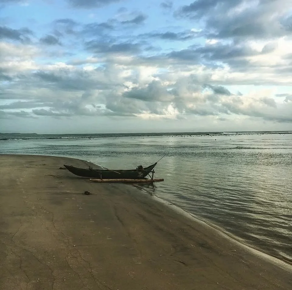 A small wooden boat on a sandy beach, with water gently lapping nearby and a cloudy sky overhead.