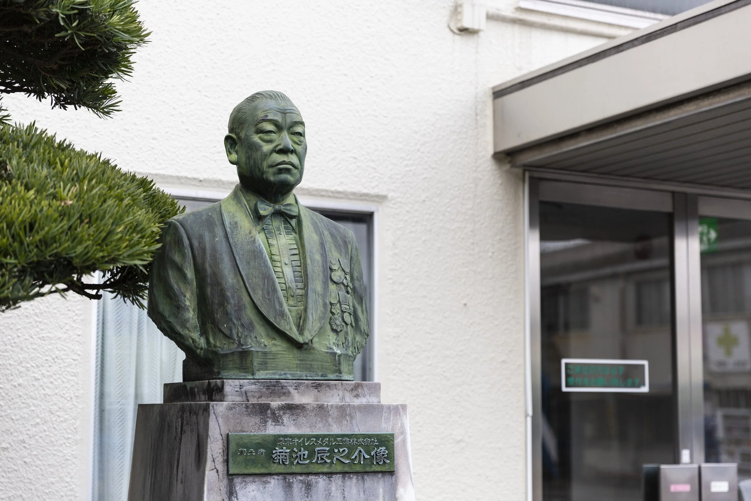 Bronze bust of a man in formal attire, including a bow tie, with a plaque in Japanese characters beneath it, placed outdoors next to bushes and a building.