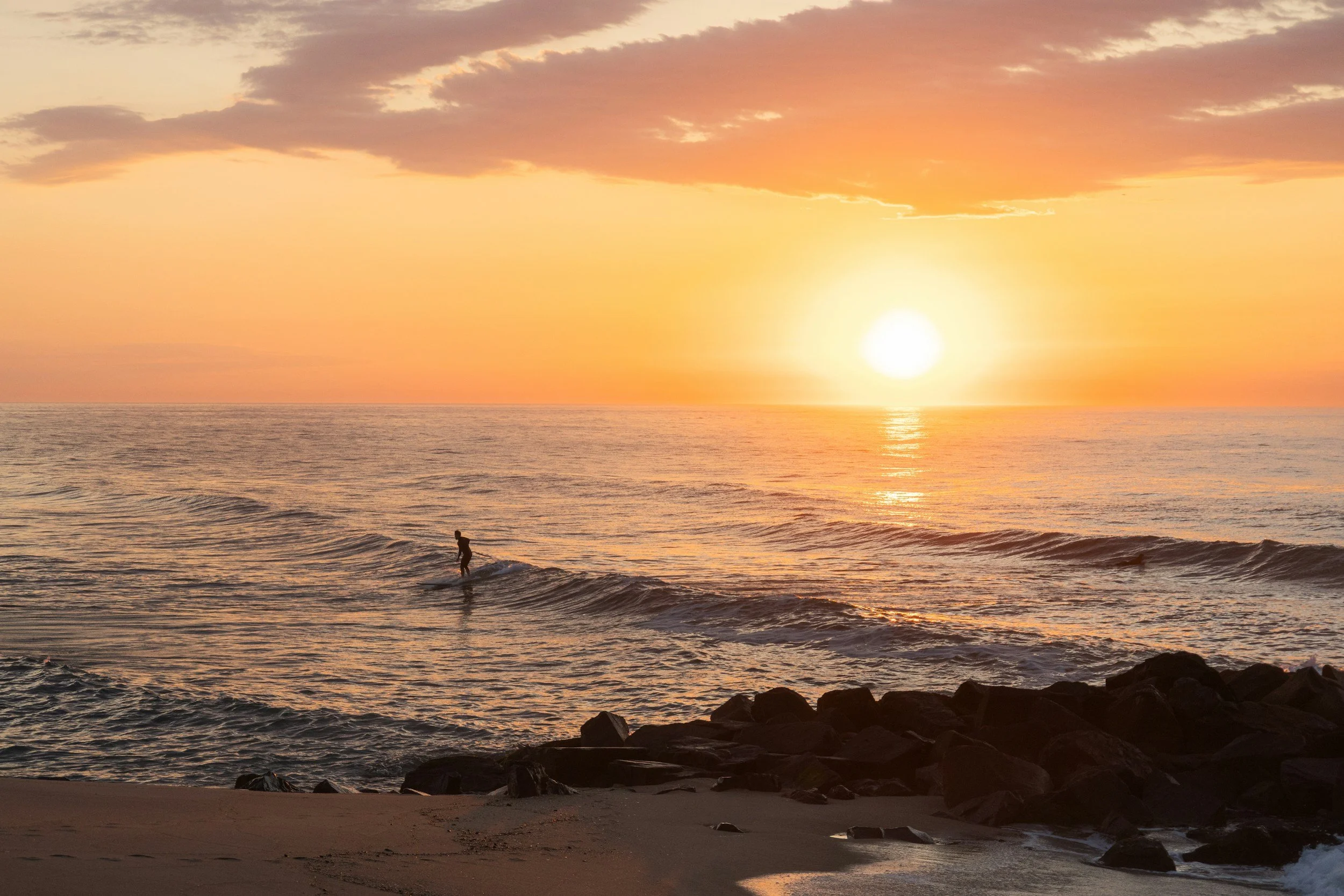 A person standing on a surfboard near the shoreline at sunset, with the sun low on the horizon and clouds in the sky.