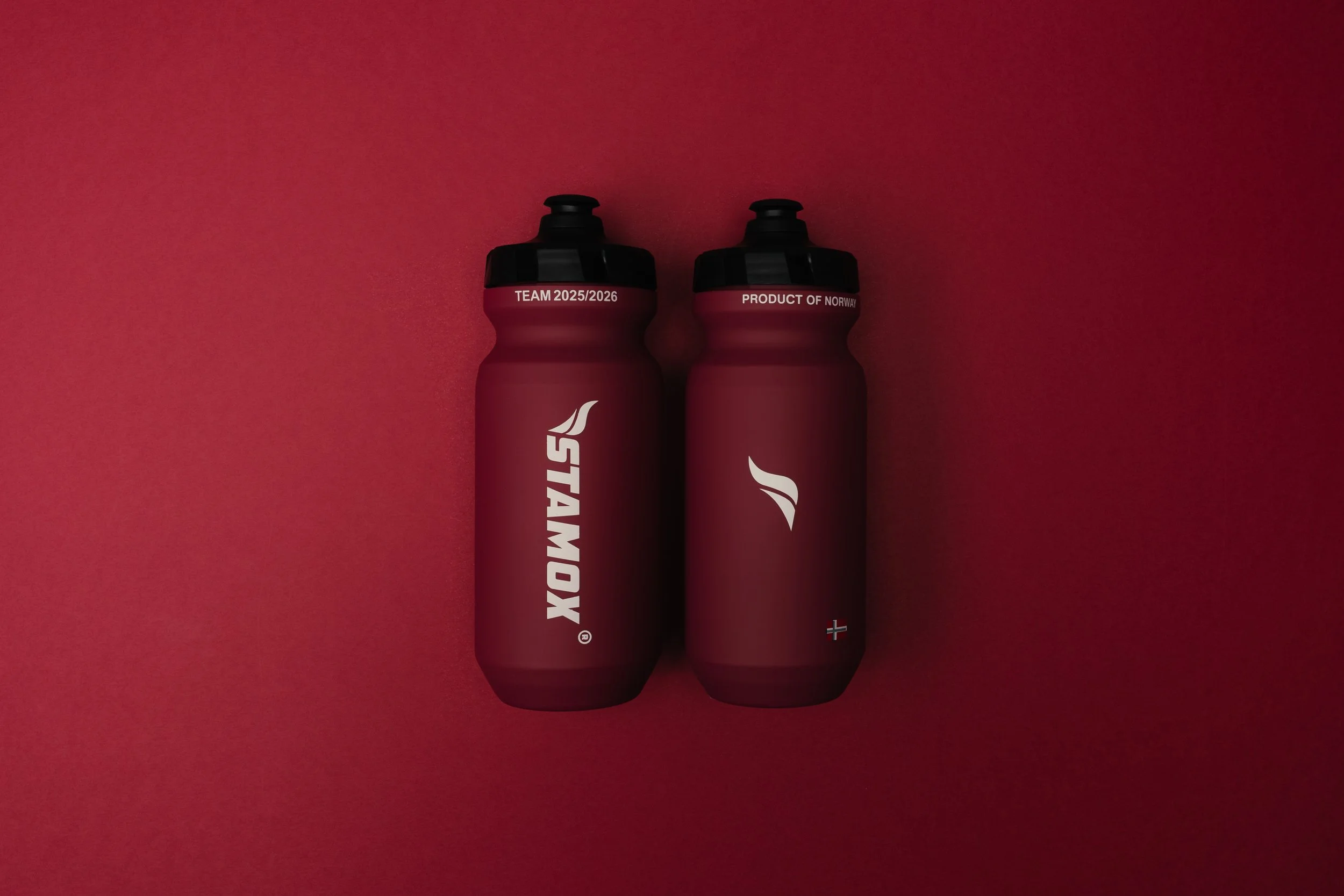 Two maroon-colored sports bottles with black caps, placed on a red background. One bottle has text "TEAM 2025/2026" and the other has "PRODUCT OF NORWAY."