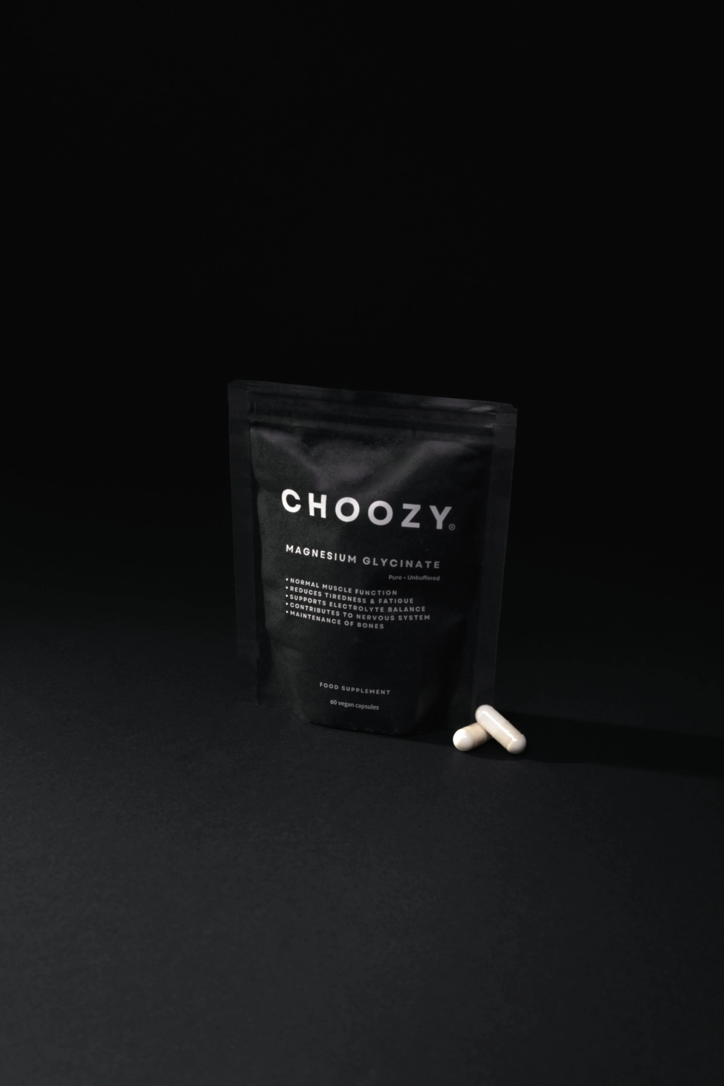 A black package labeled 'CHOOZY Magnesium Glycinate' standing upright on a black surface, with two white capsules placed in front of it. The package contains 60 vegan capsules and lists benefits like supporting nerve system and maintaining bones.
