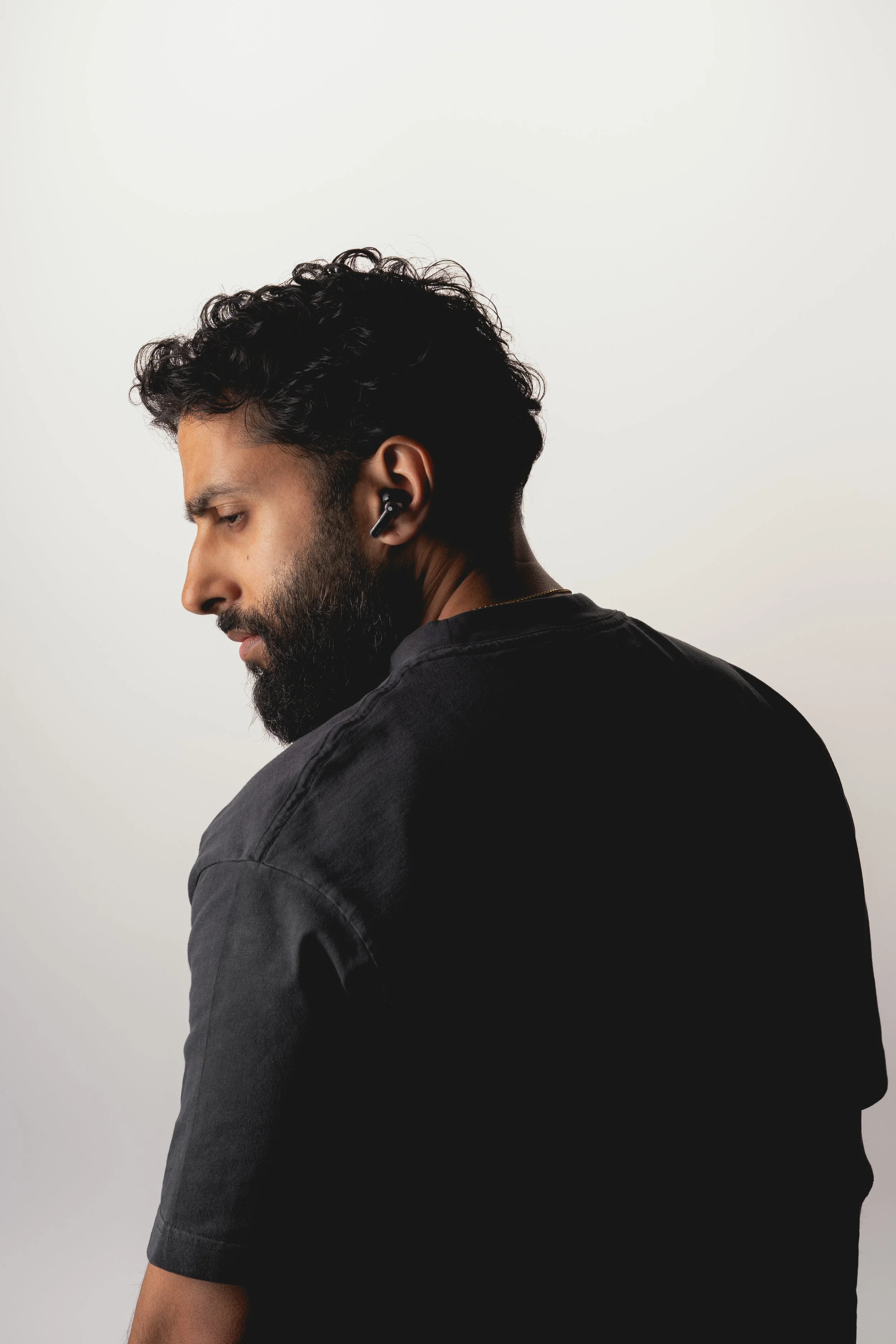 A man with dark, curly hair and a beard is wearing wireless earbuds and a black shirt, looking down with a neutral expression against a plain white background.