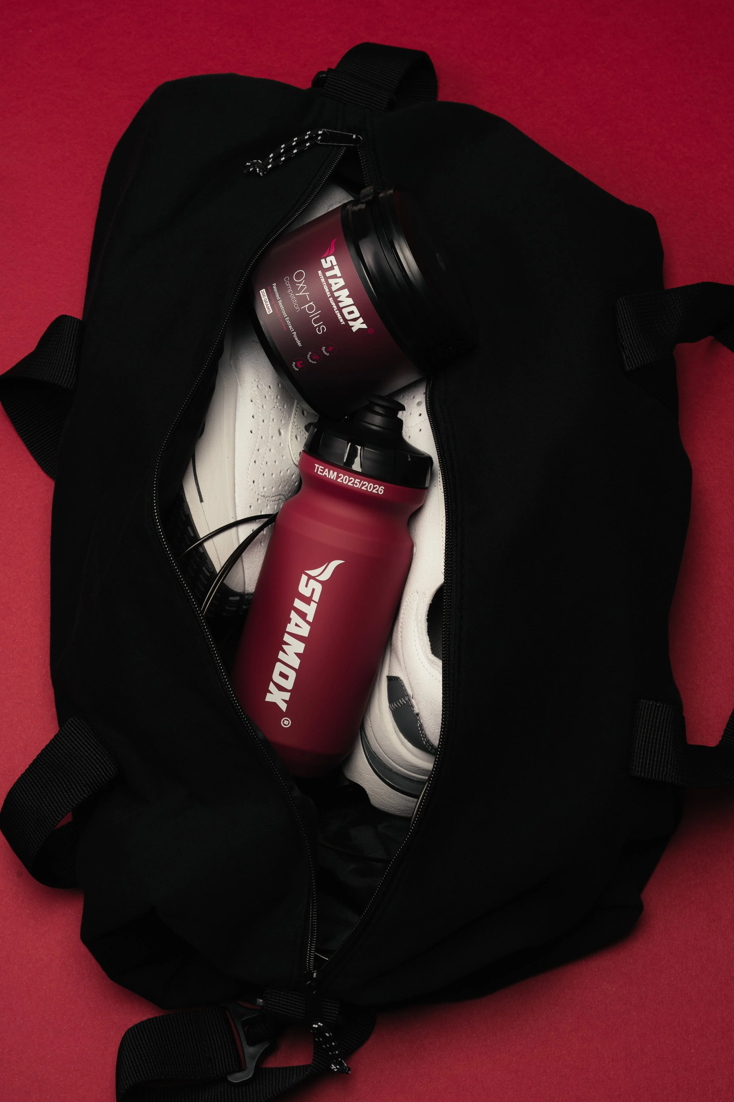 Black sports bag open with a red and black stainless steel water bottle and a container of Oxylite Plus supplement inside, on a red surface.