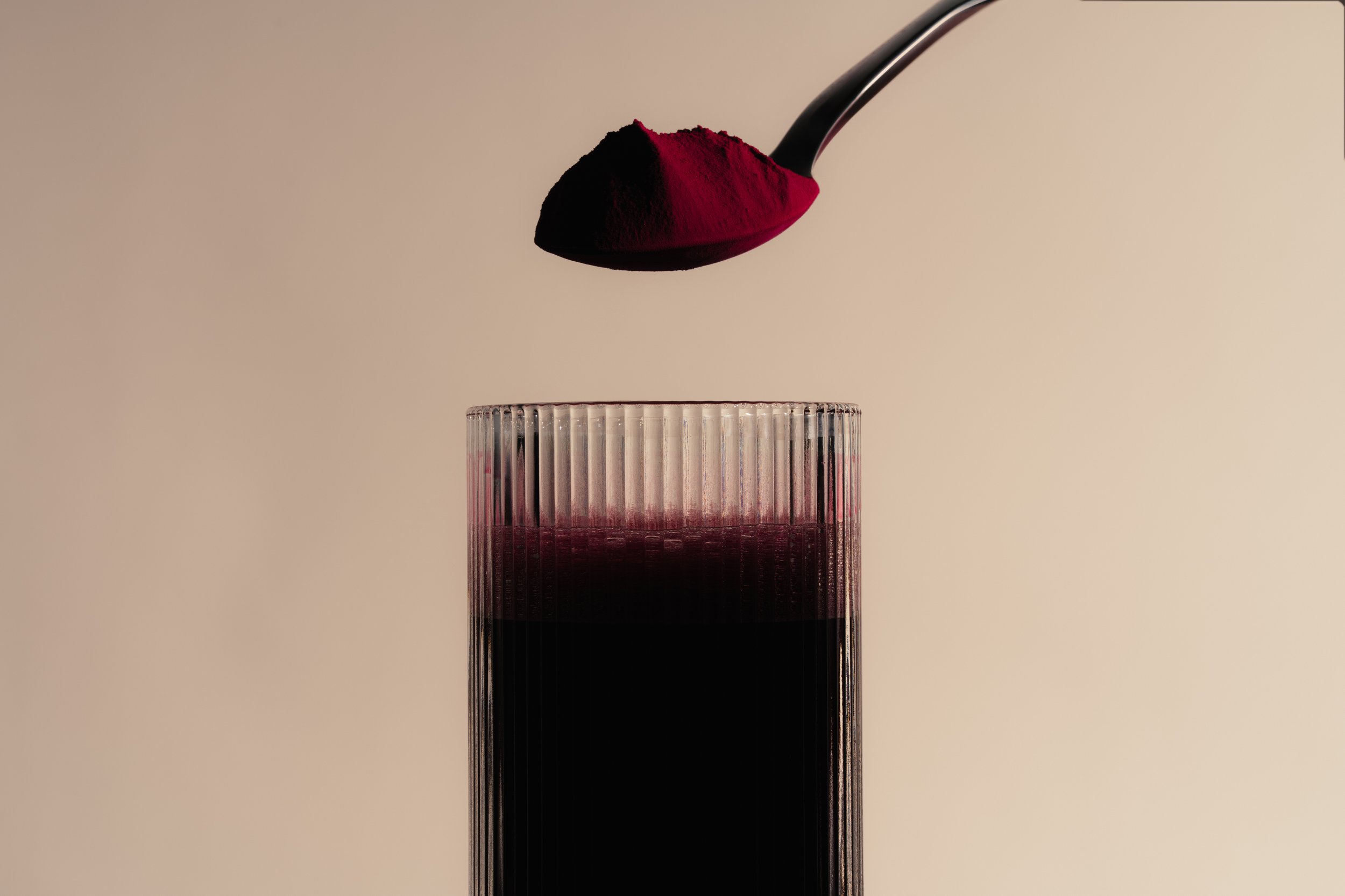 A spoon with a reddish-fuchsia powder above a textured glass filled with dark liquid.