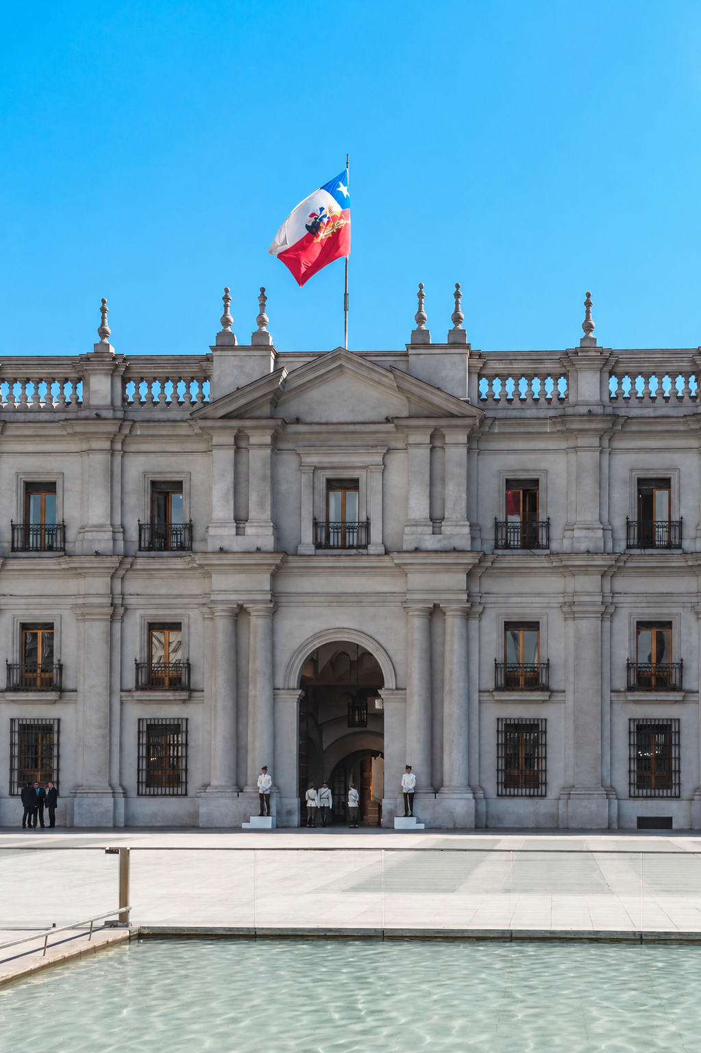 Edificio gubernamental con bandera de Chile ondeando en el techo, fachada de piedra, ventanas con barandillas y guardias en la entrada, frente a una fuente y un cielo azul claro.