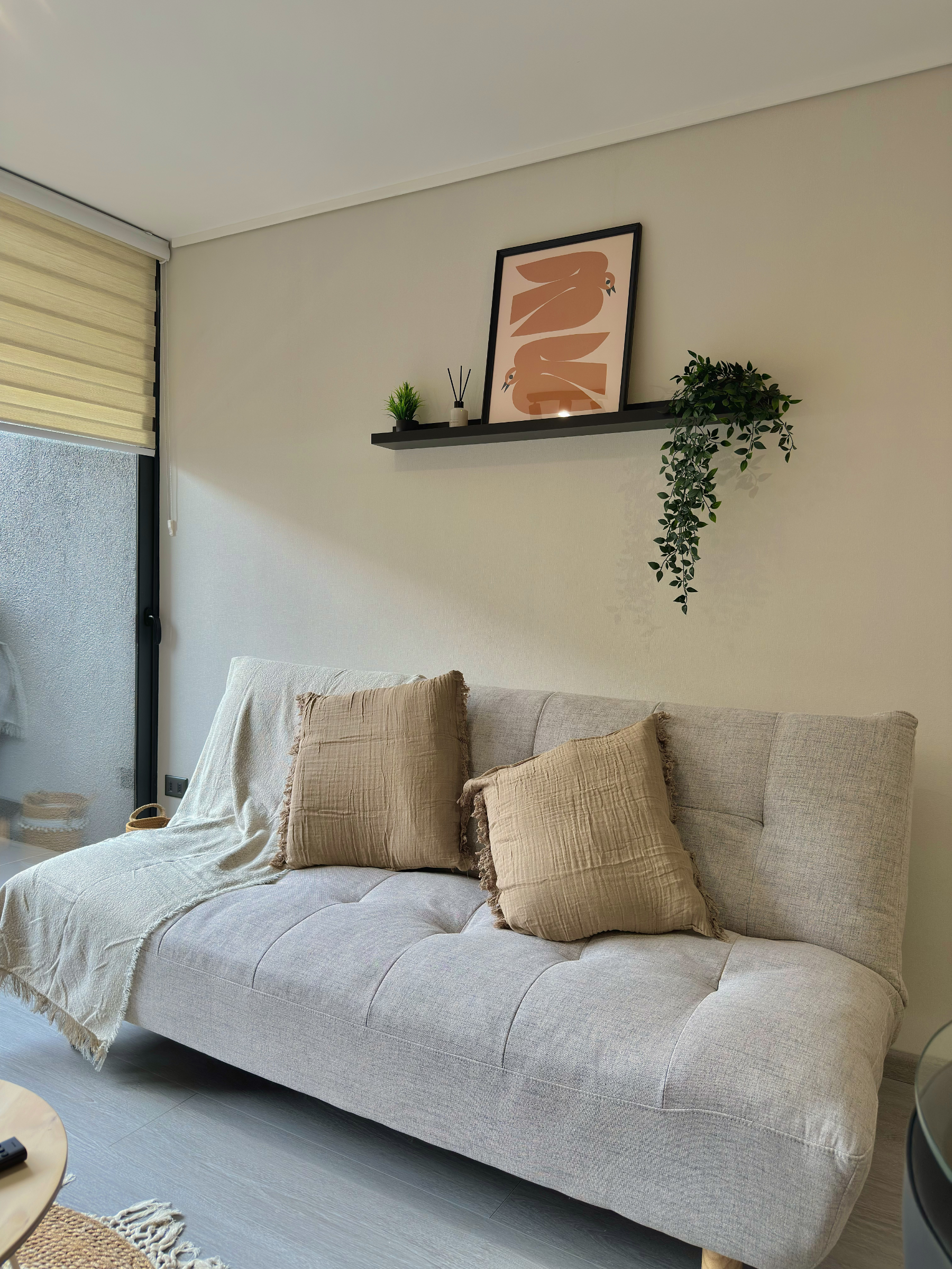 Sofá cma gris con cojines beige, en una sala con ventana con persiana beige y una repisa con plantas y cartel decorativo