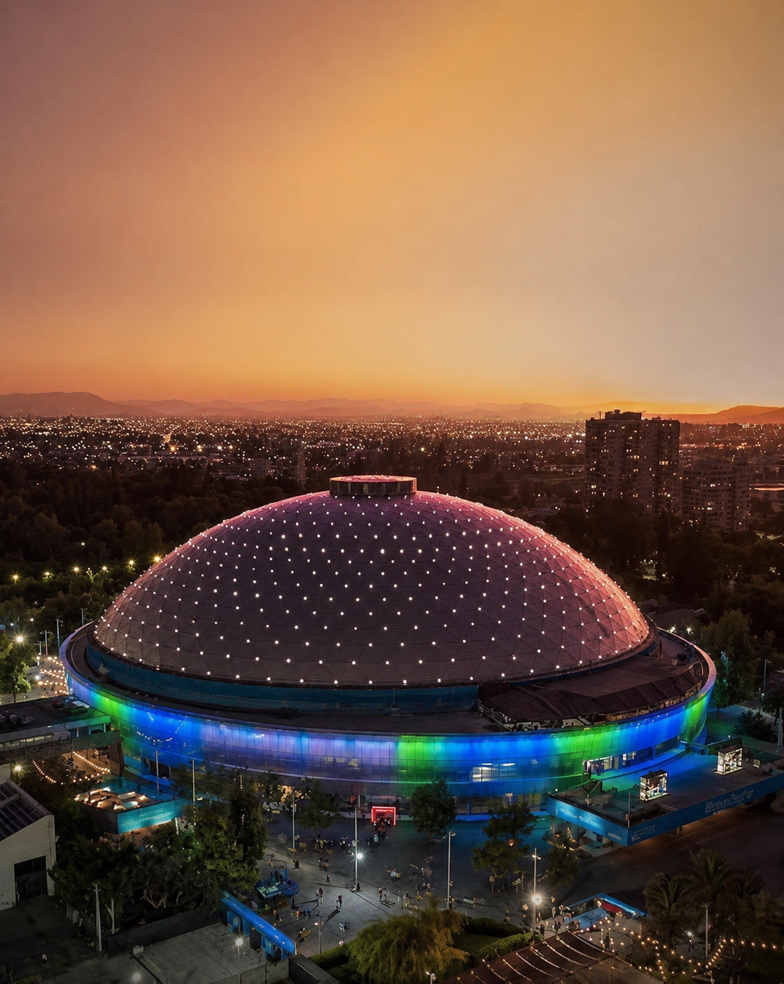 Vista aérea del Movistar Arena en Santiago con luces de colores en un atardecer en la ciudad cercano a uno de los departamentos.