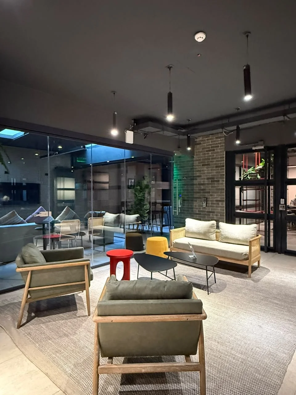 Sala de espera moderna con sillones, mesas y plantas, vista a un área cerrada con ventanas de vidrio y decoración acogedora.