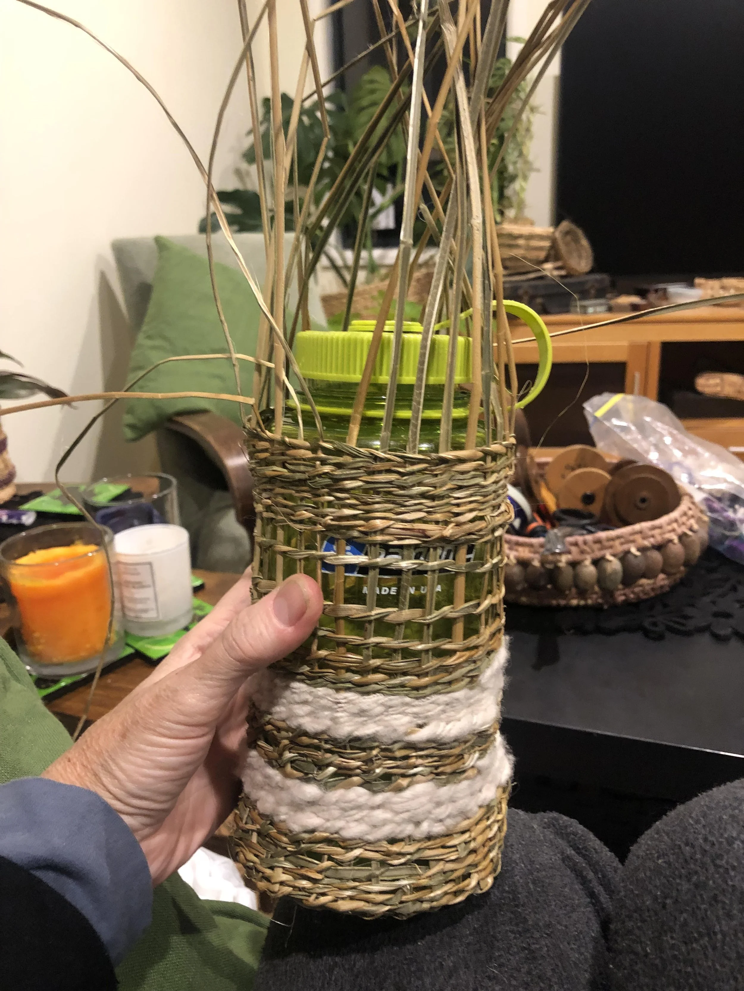 Close-up of the makings of a twined basket with a green water bottle inside, held by a hand. The background features candles, a green pillow, and various items on a table and shelf in a cozy indoor setting.