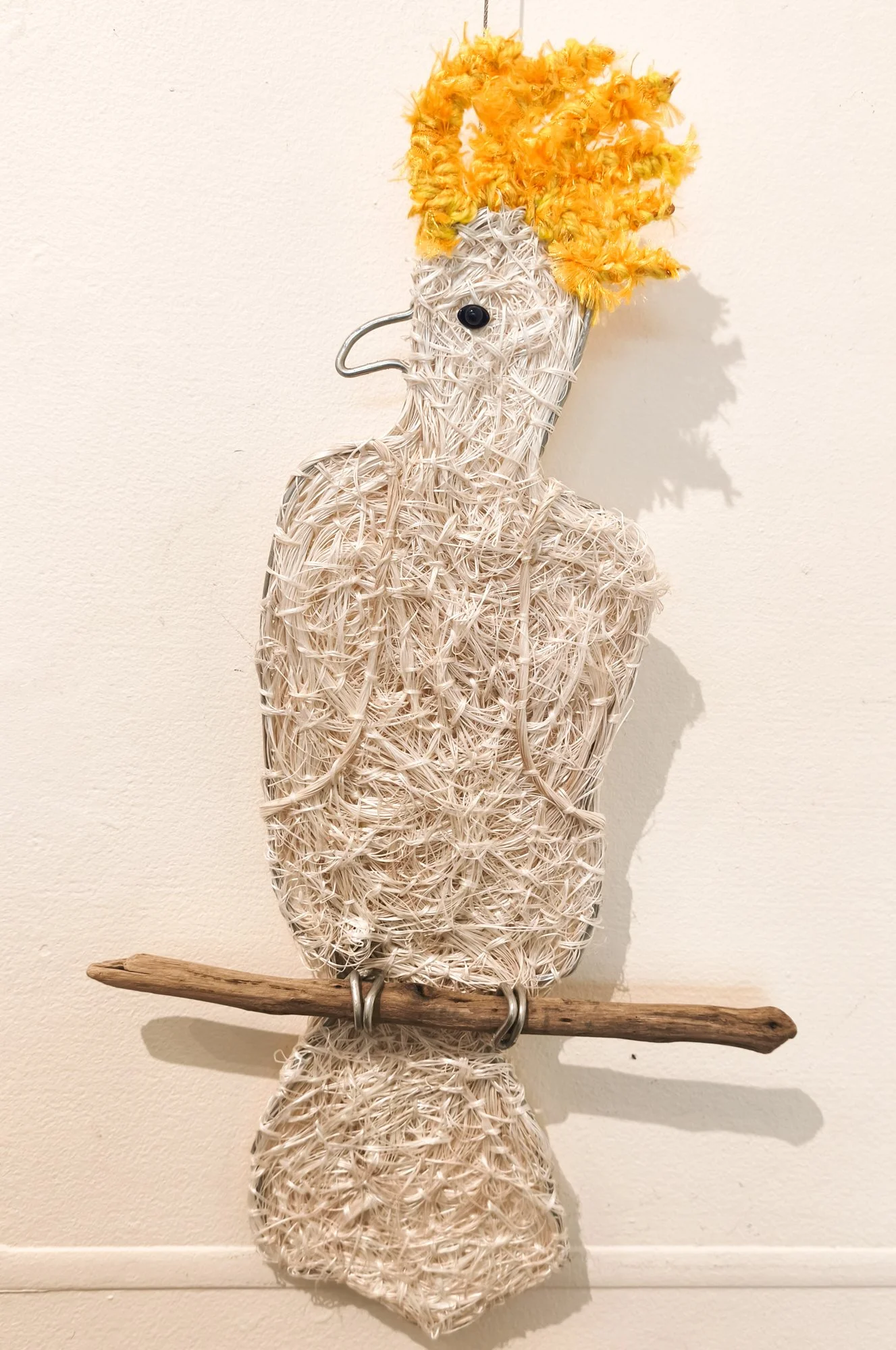 A flat fibre sculpture of a Cockatoo with yellow crest, made of recycled rope material, holding a wooden stick, mounted on a wall.