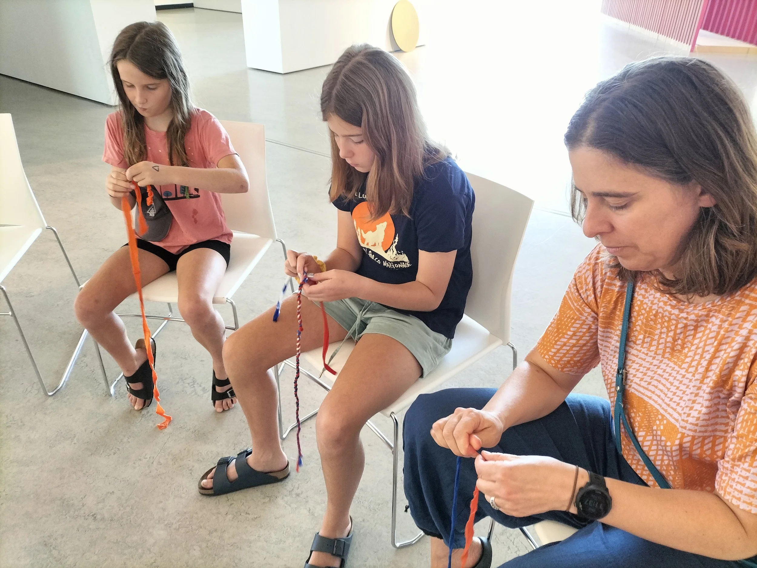 Three children sitting in chairs, tying colorful ropes, indoors with a white wall and some decorative elements in the background.