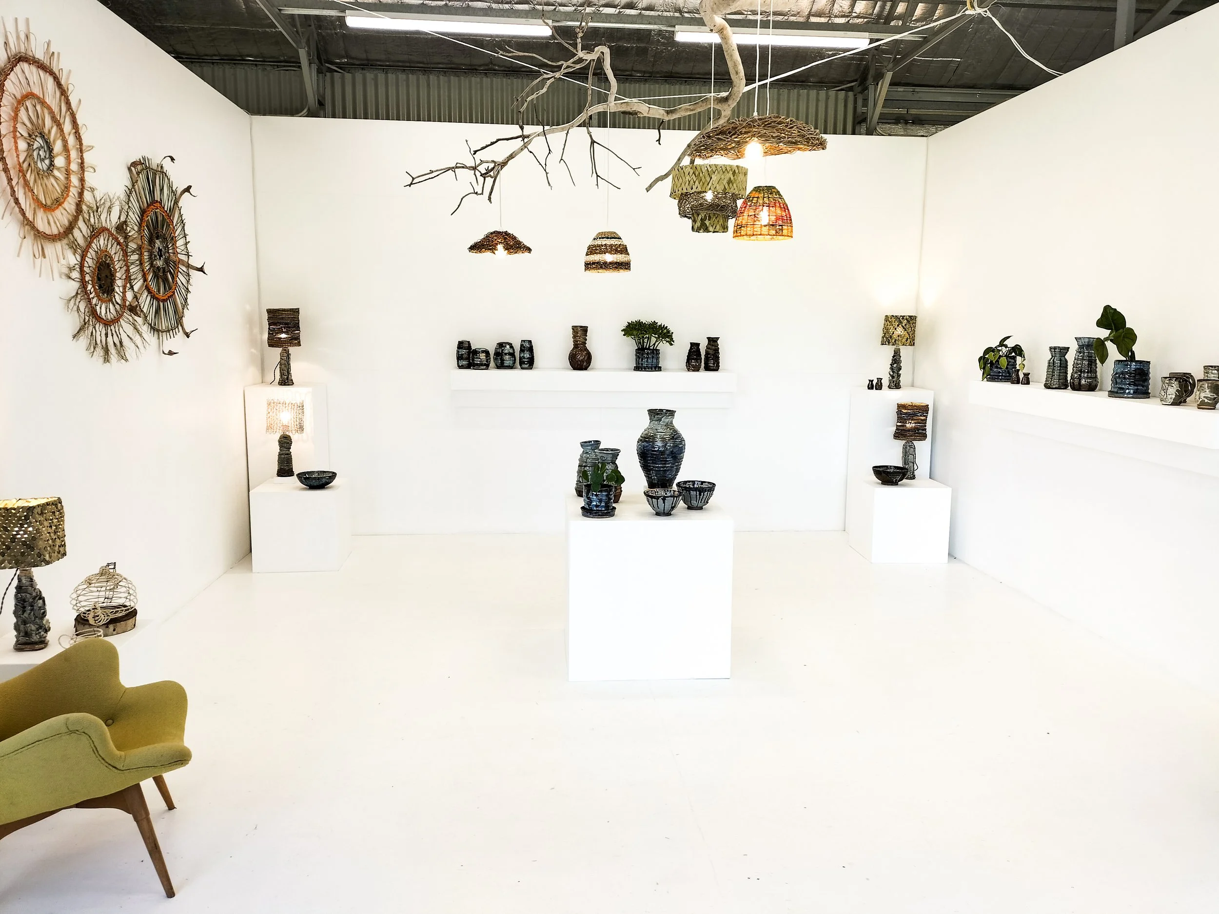 Display of ceramic and woven art pieces on white walls and shelves in a gallery, with decorative lamps and a green armchair in the foreground.