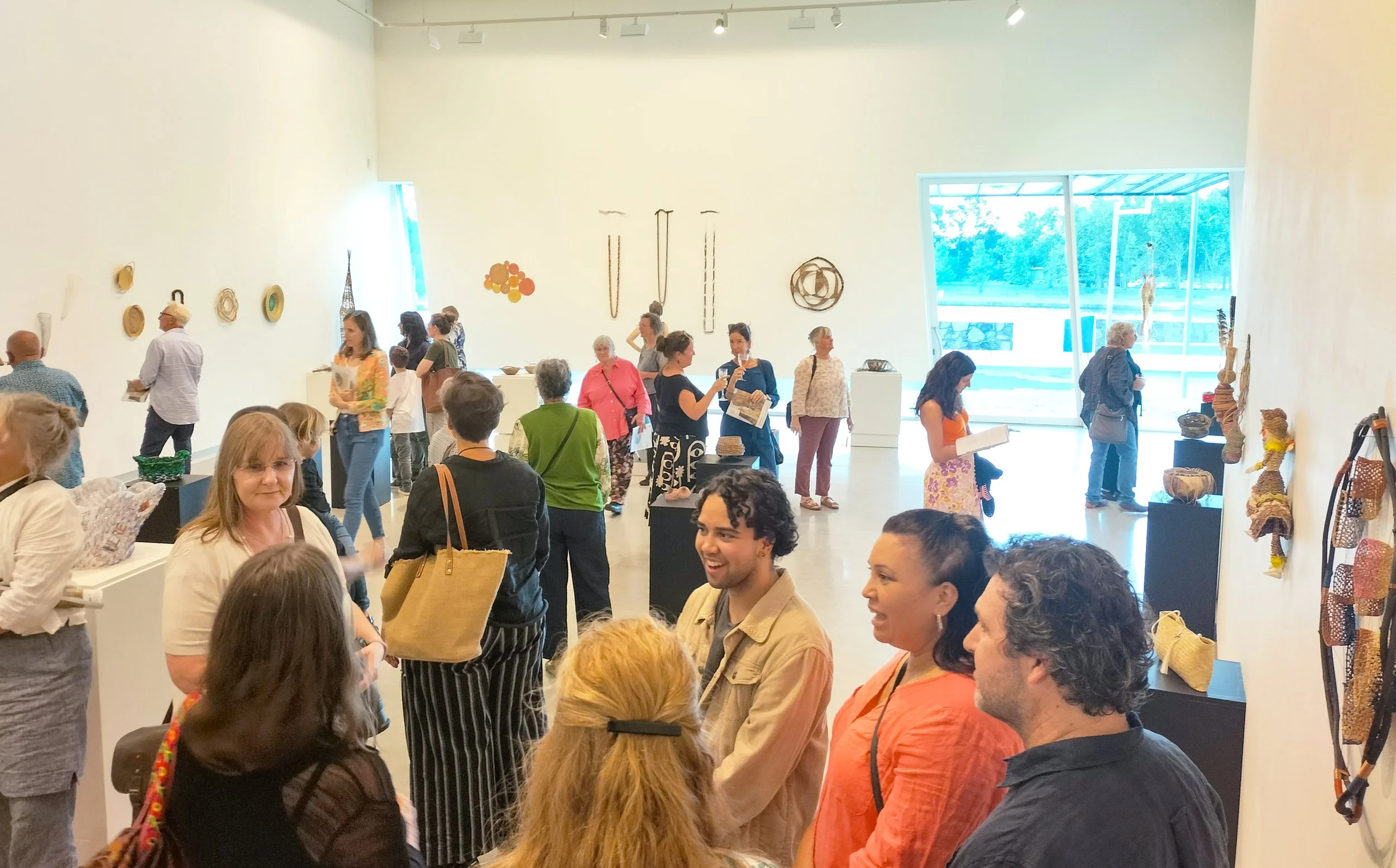 People attending an art exhibition in a bright, modern gallery with white walls and high ceilings, displaying various sculptures and woven art pieces.