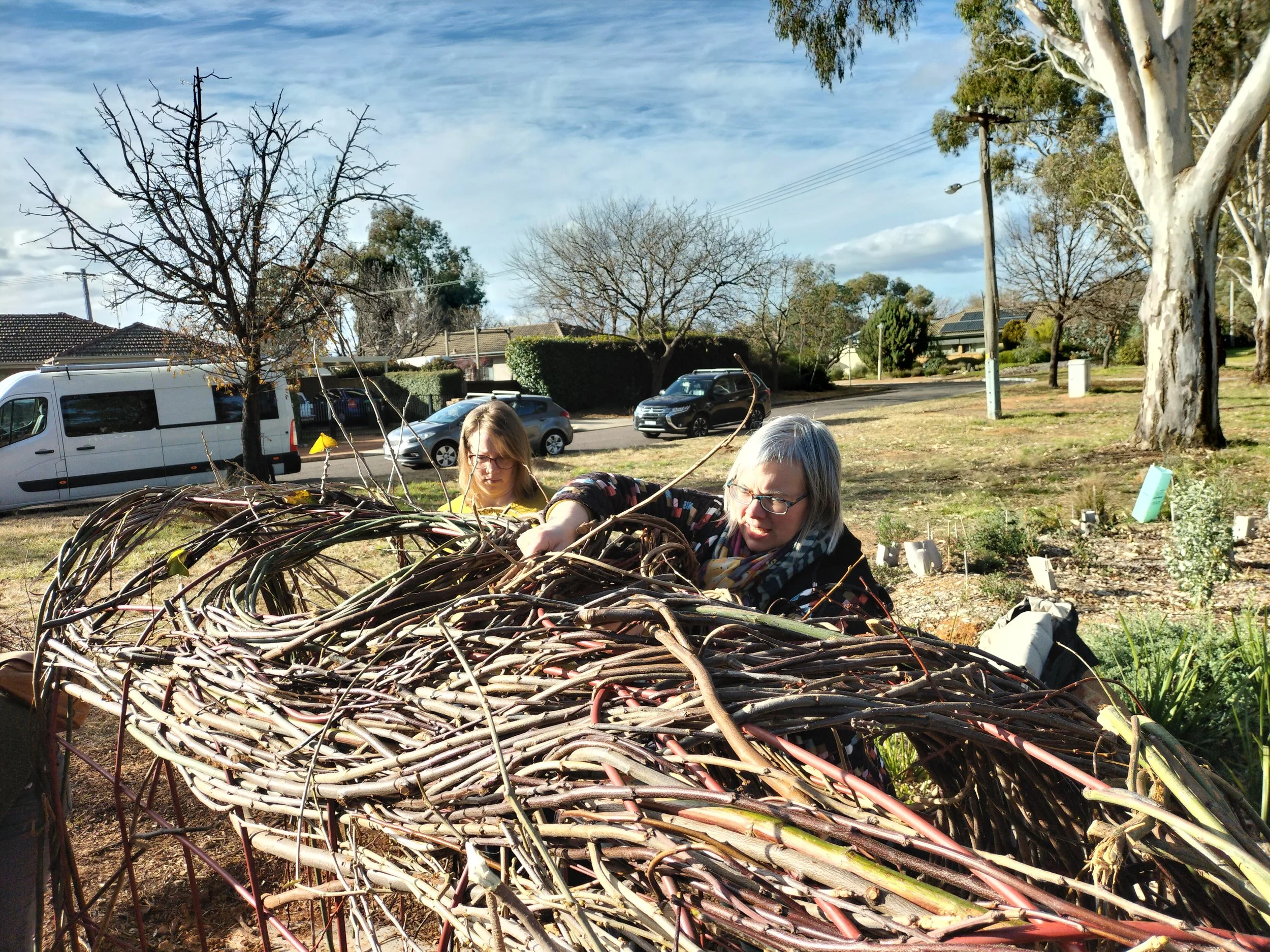 Two women working together on a large woven basket made from intertwined sticks outdoors in a yard with trees and parked cars.